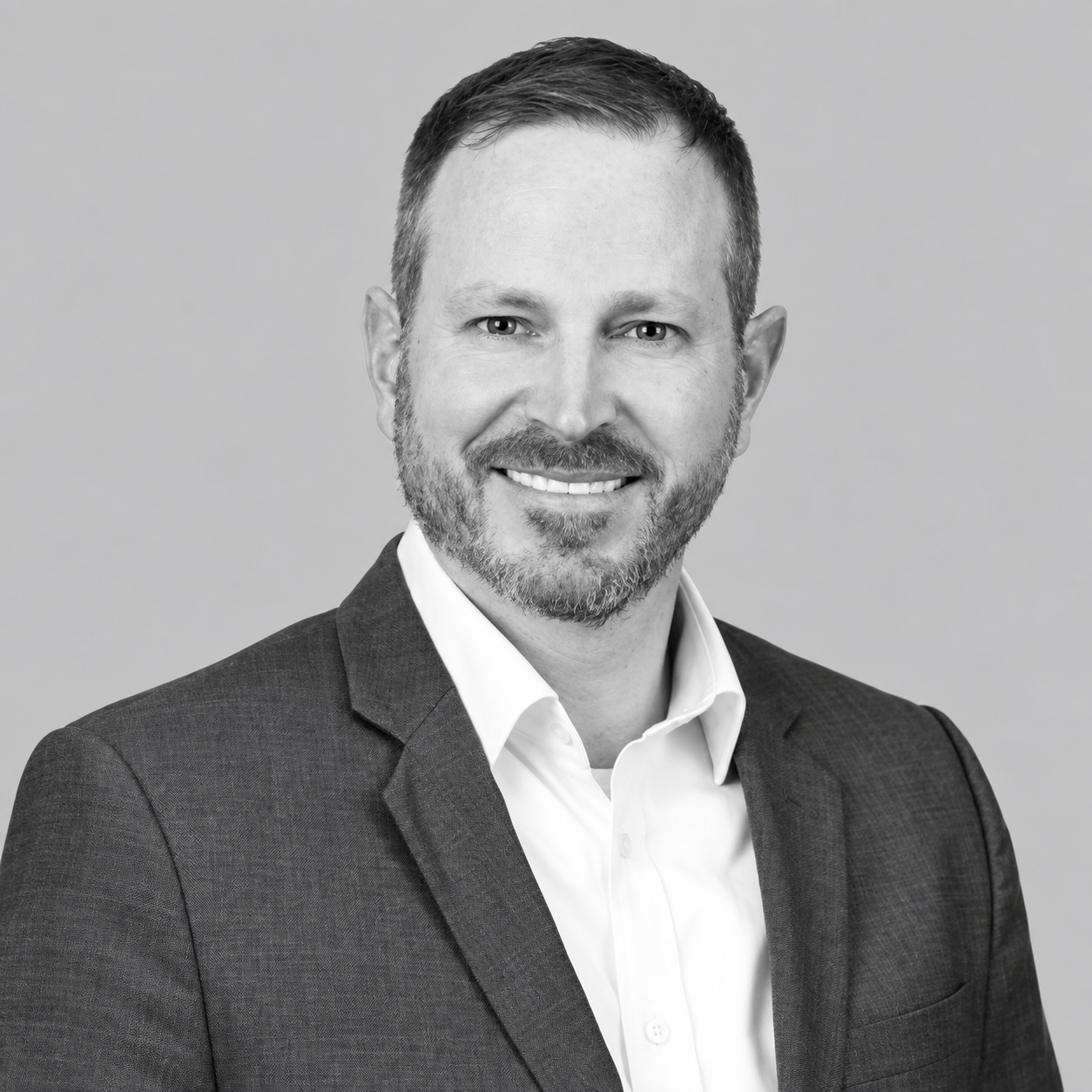 Michael Gietl -Black and white headshot of a smiling man with short hair and a beard, wearing a suit jacket and white shirt.