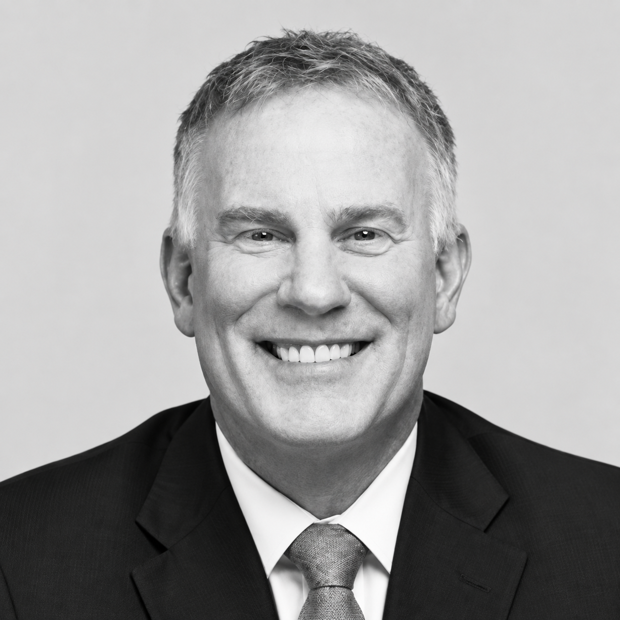 Pat Mascia - Black and white portrait of a smiling middle-aged man with short, light hair, wearing a dark suit, white shirt, and patterned tie.