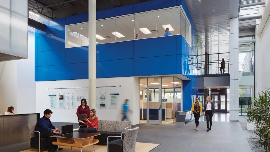 Interior of a modern building with a two-story glass and blue panel structure, people walking and sitting, and a reception area.