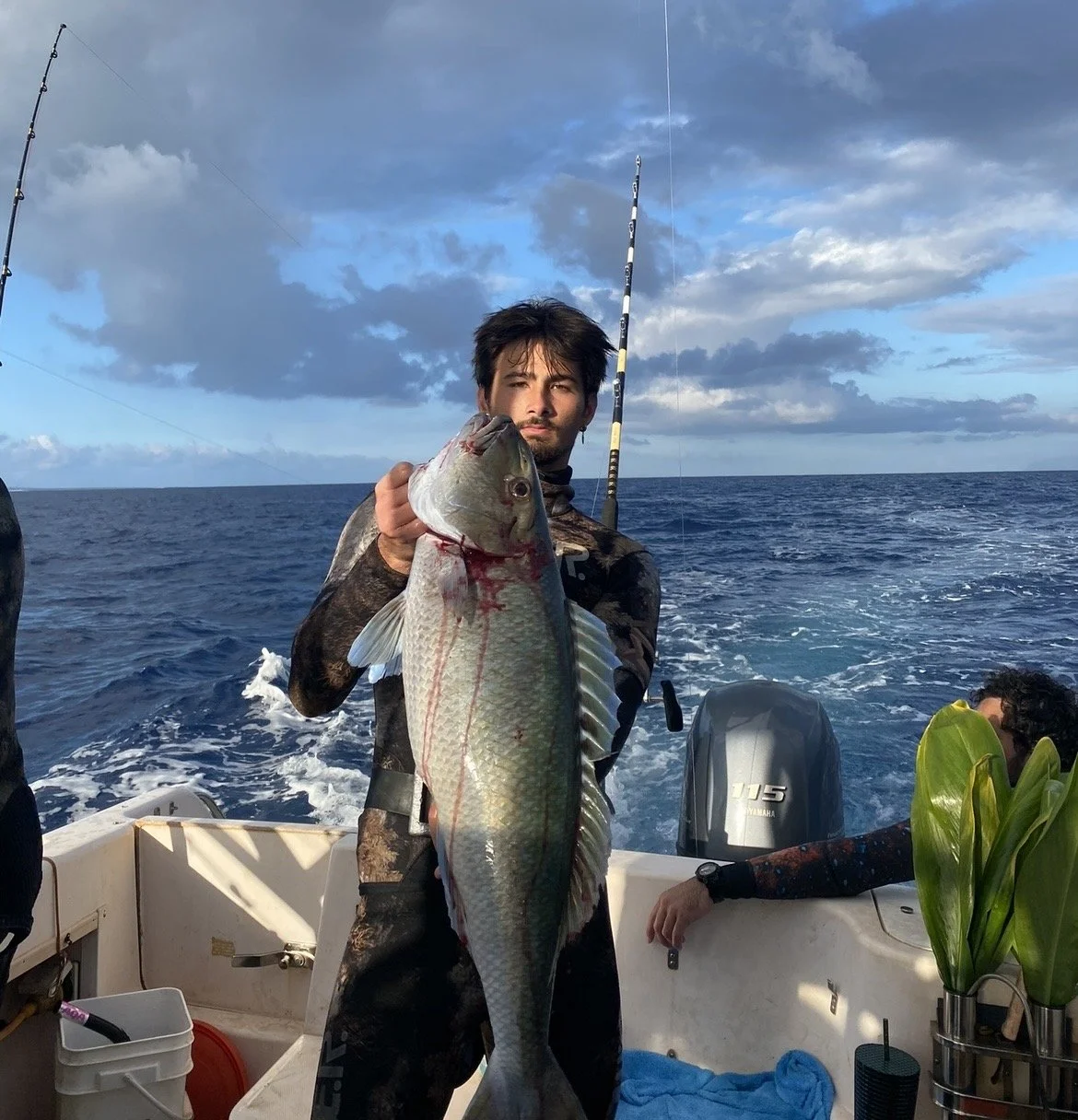 A man holding a large fish on a boat in the ocean, with fishing rods in the background and cloudy sky overhead.