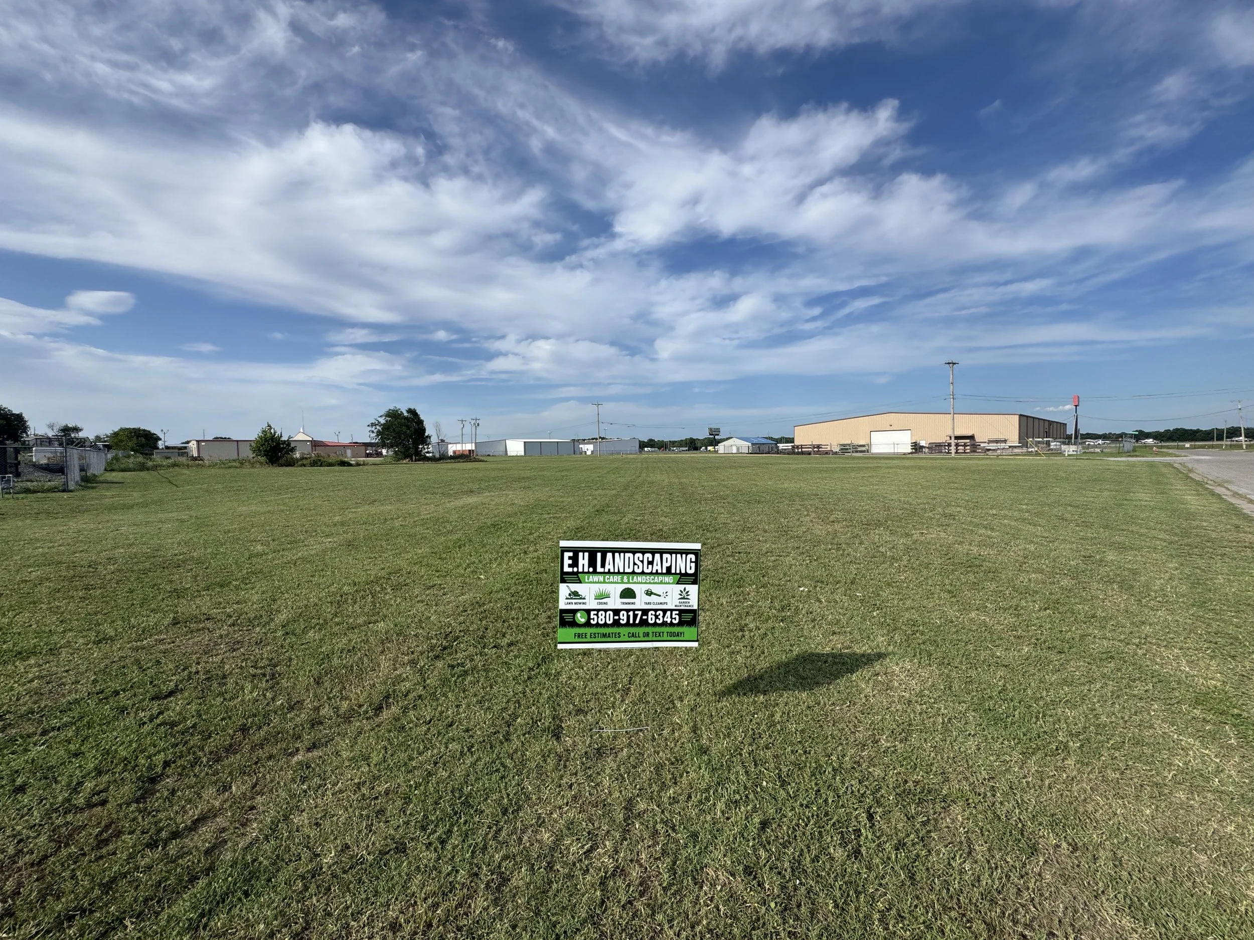 A vacant grassy lot with a sign advertising landscaping services, set against a backdrop of industrial buildings and a partly cloudy sky.