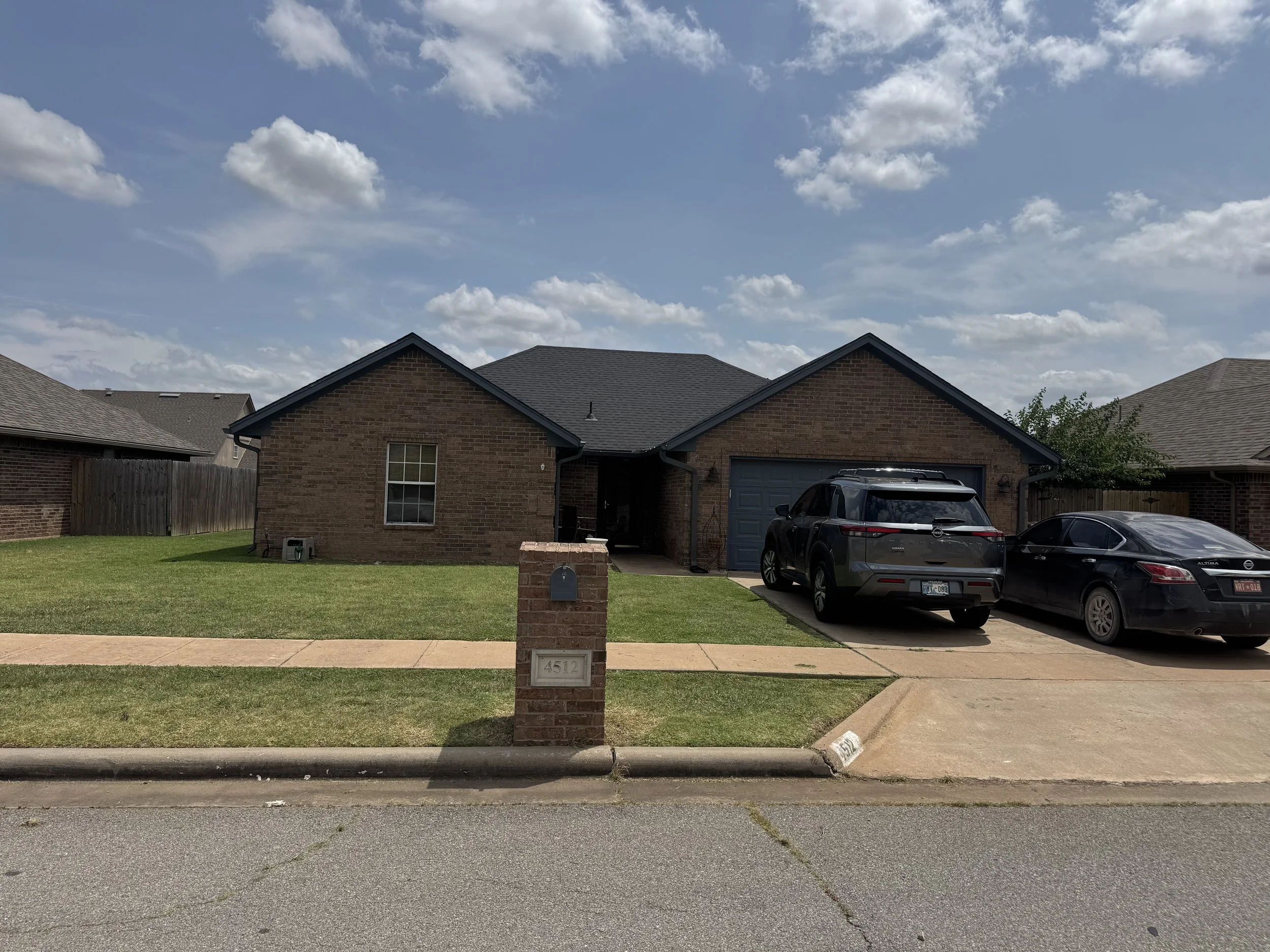 View of a brick house with a black roof, a blue garage door, and a driveway with two parked cars, a mailbox with the number 4512, a sidewalk, and a green lawn under a partly cloudy sky.