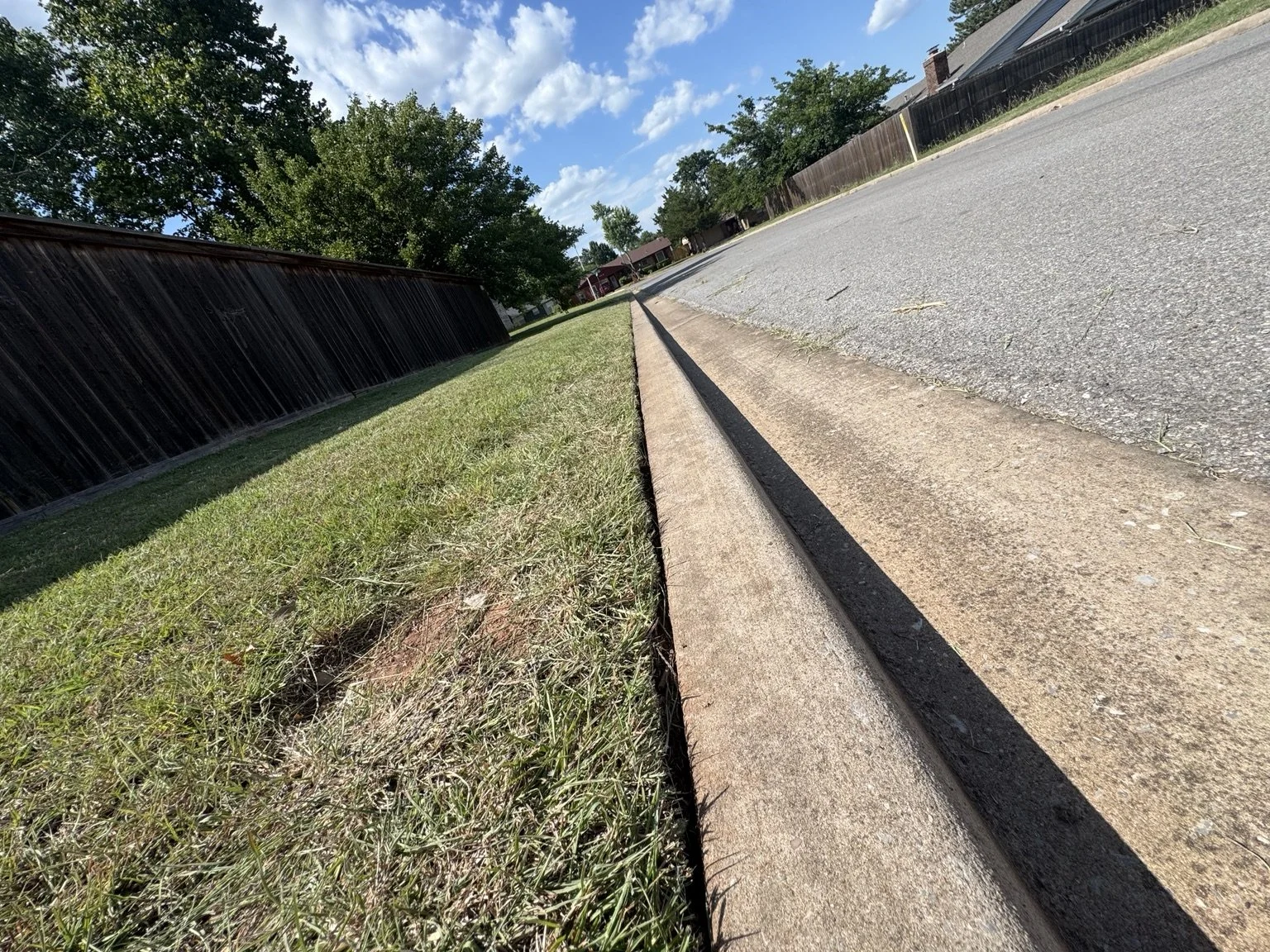 Sidewalk and street view with a wooden fence, trees, and houses in the background under a blue sky with clouds.
