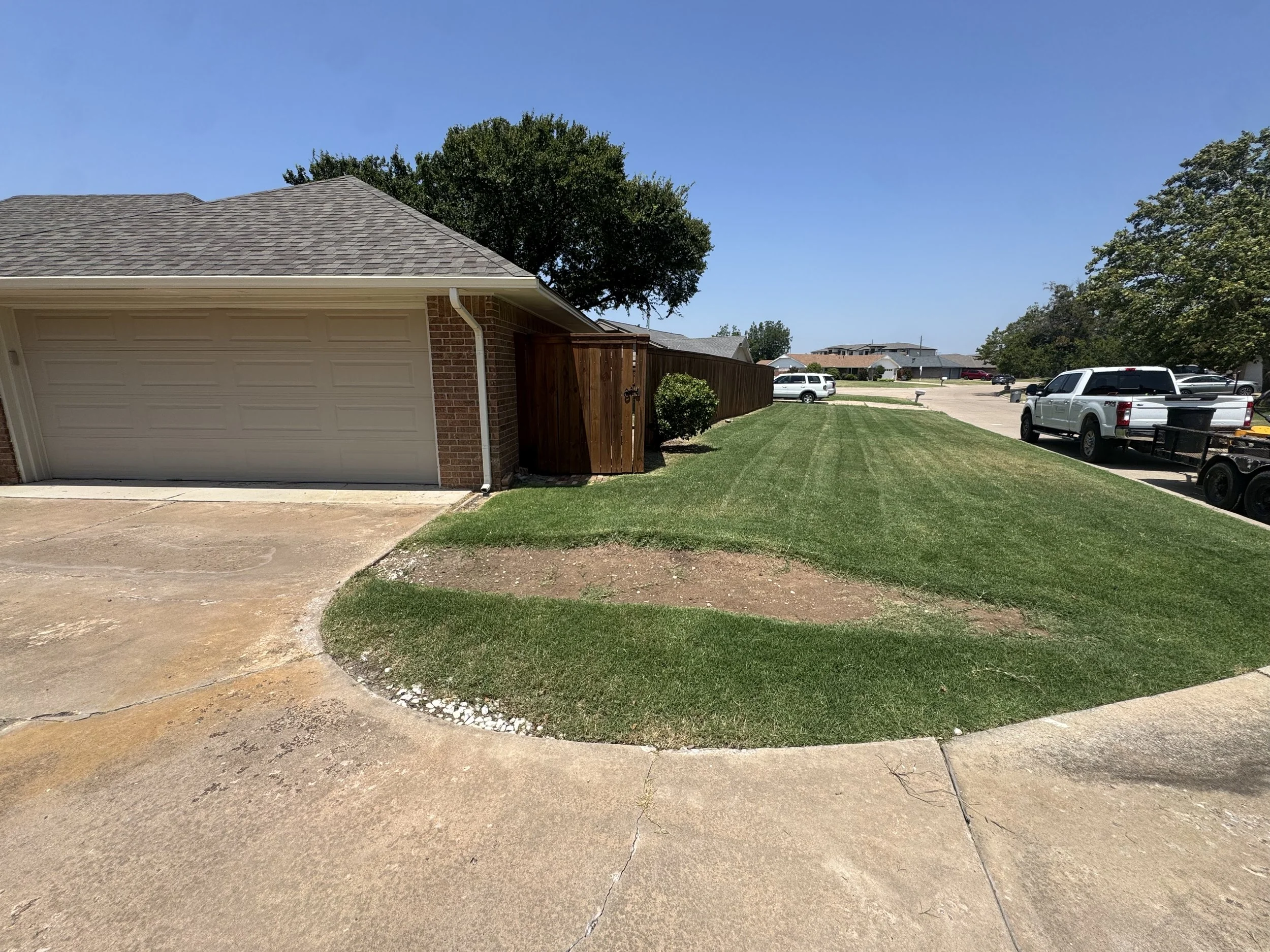 A suburban neighborhood yard with a grass lawn, a sidewalk, parked cars, trees, and houses under a clear blue sky.