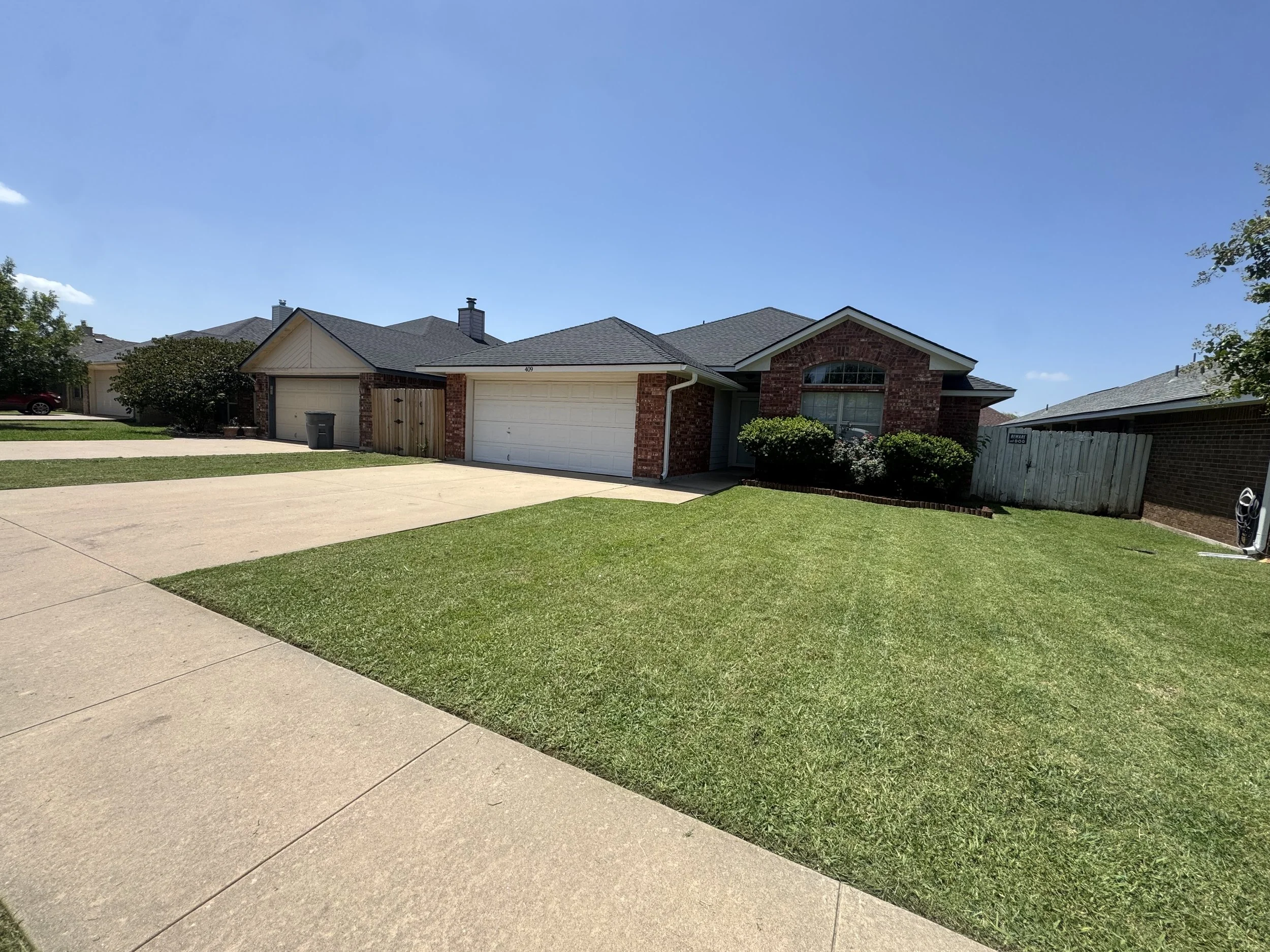 A suburban house with a brick exterior, a garage, well-maintained lawn, and a sidewalk in front, under a clear blue sky.