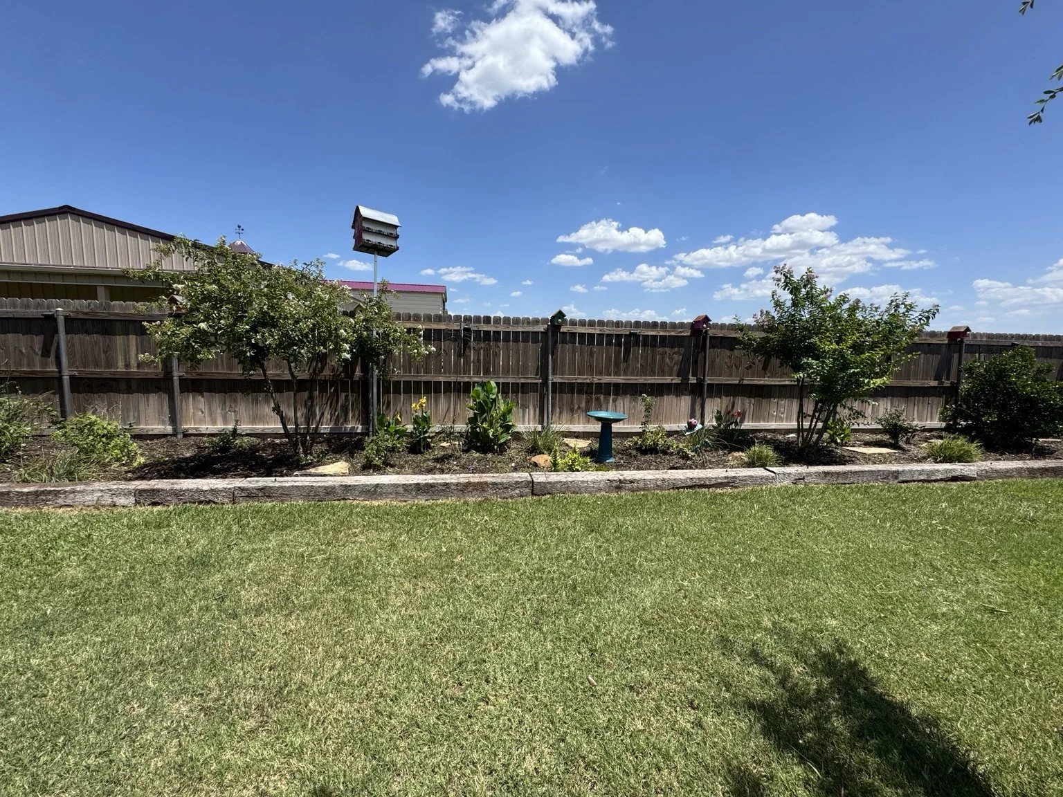Backyard garden with a wooden fence, two trees, flower bed, bird bath, and a lawn under a blue sky with clouds.