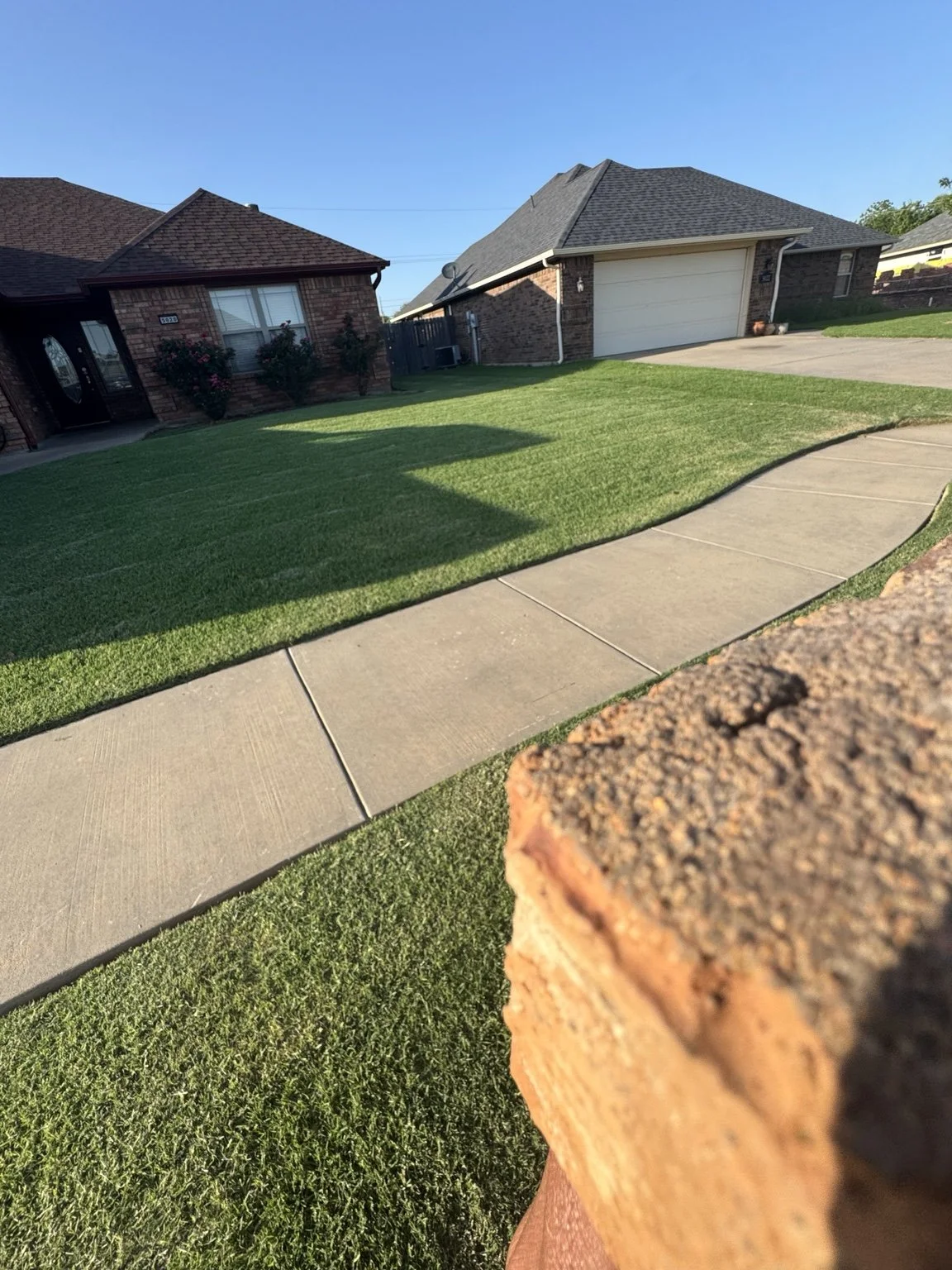 A suburban house with a brick exterior, well-manicured lawn, concrete sidewalk, and a driveway leading to a closed garage, under a clear blue sky.