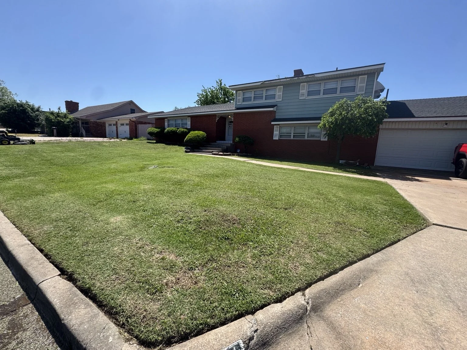 A residential front yard with green grass, trimmed bushes, a concrete walkway leading to a front entrance, and a two-story house with brick and siding exterior. A tree is on the right side, and a driveway with a parked vehicle is on the right.