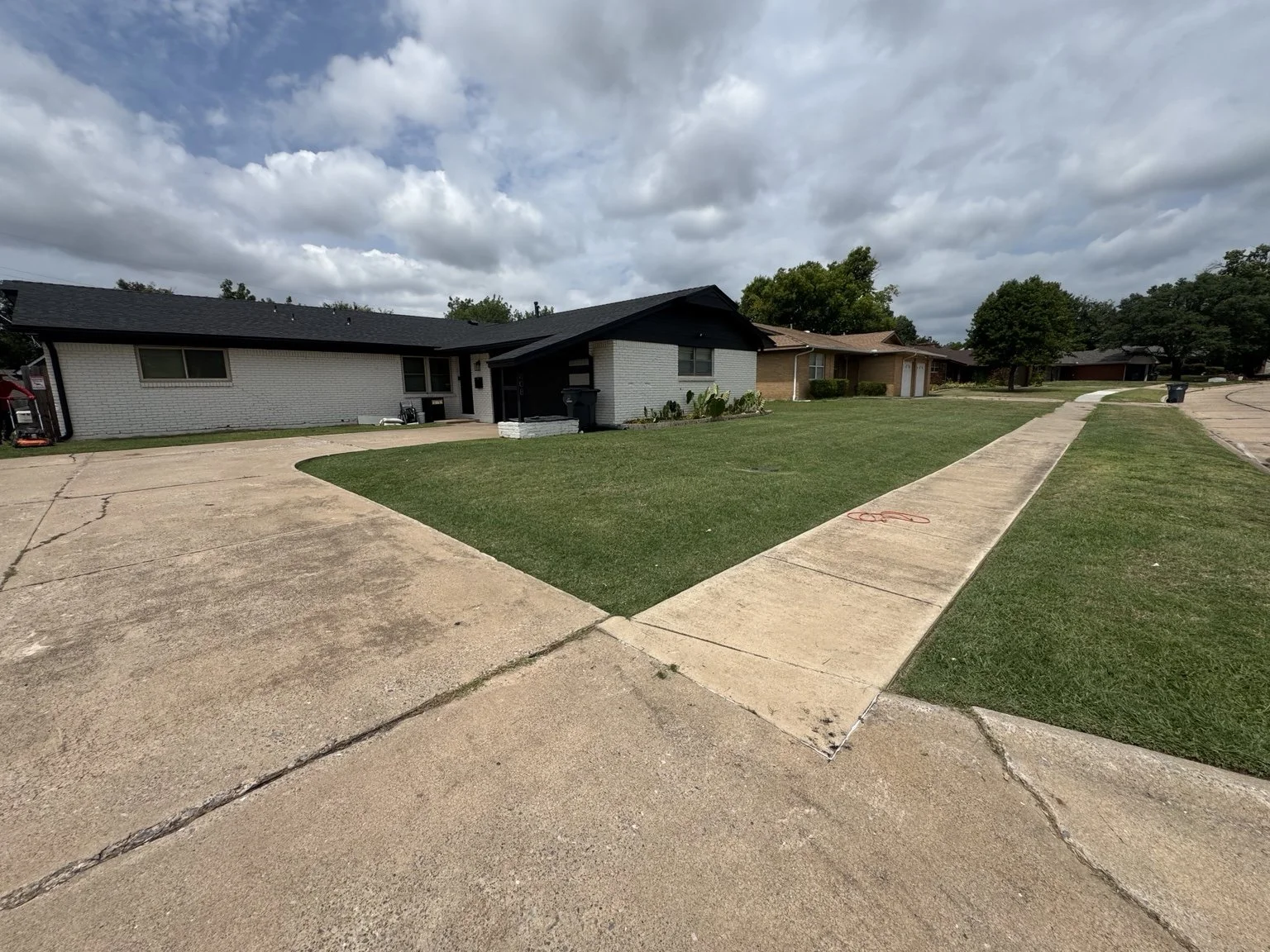 Residential neighborhood with single-story houses, a green lawn, concrete sidewalks, and a cloudy sky.