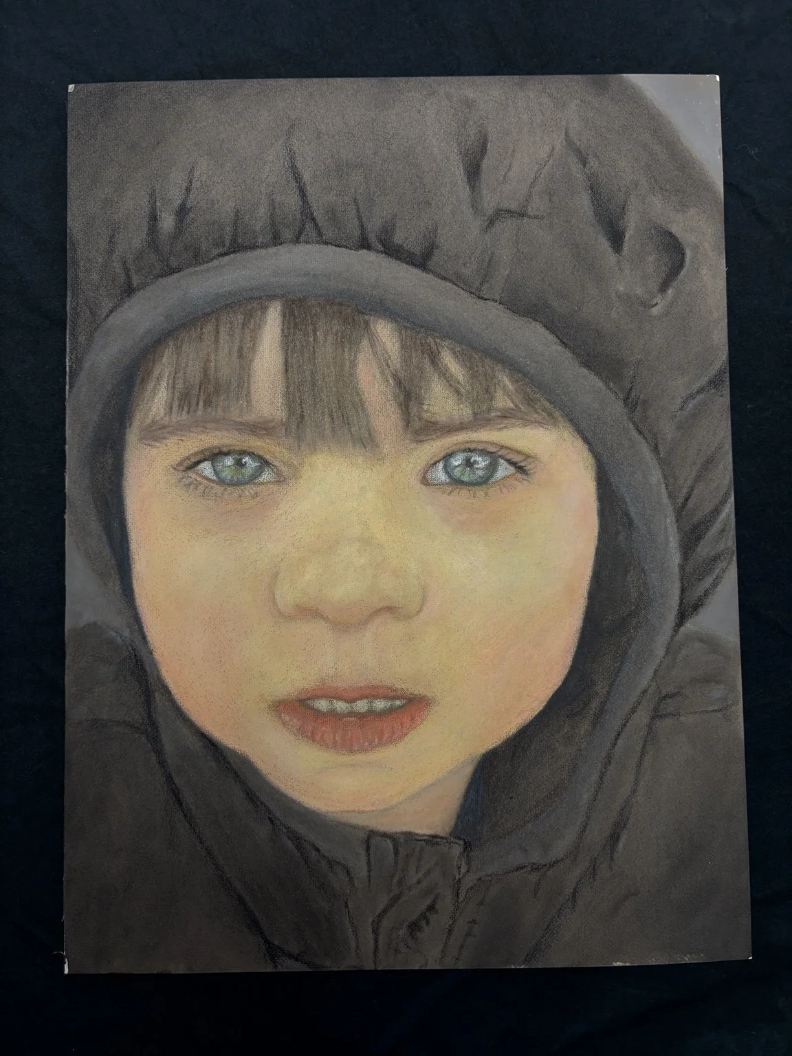A realistic colored pencil drawing of a young child's face wearing a hooded jacket with the hood up, with striking blue-green eyes, light skin, and dark hair with bangs.