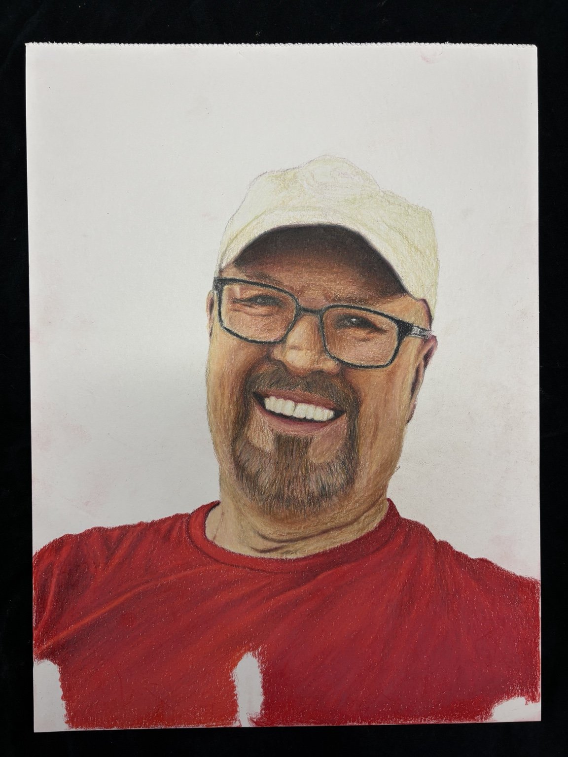 Colored pencil drawing of a smiling man wearing glasses, a white cap, and a red shirt.