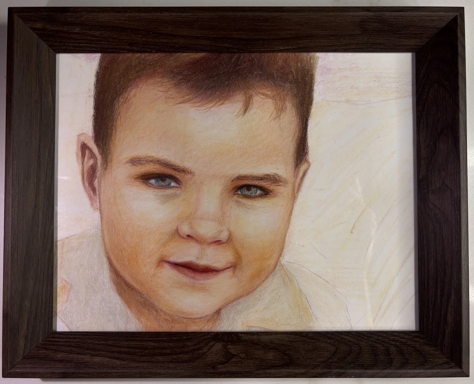A framed colored pencil portrait of a young boy with short brown hair, blue eyes, and a slight smile, against a light background.