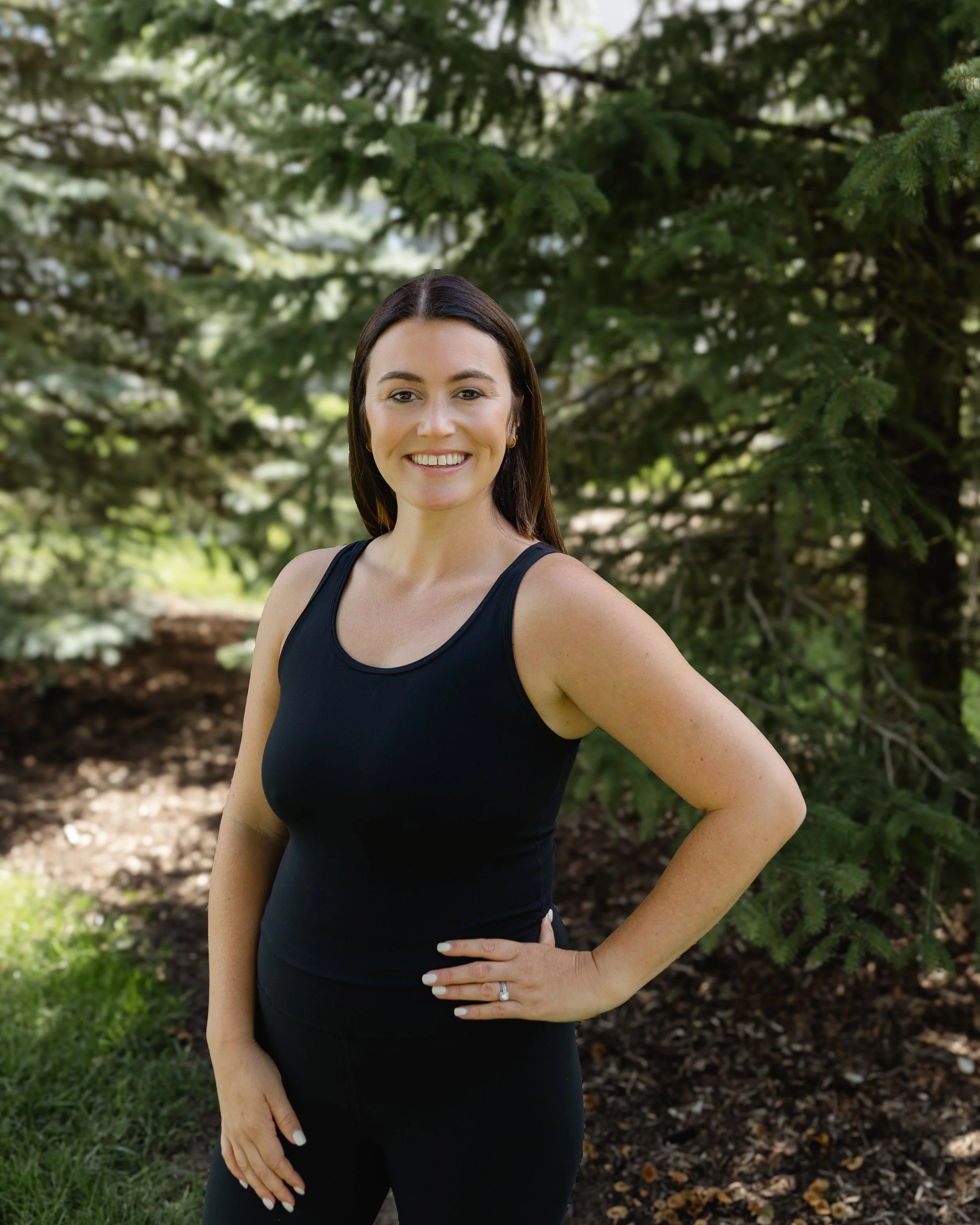A smiling woman with long brown hair standing outdoors near green trees, wearing a black tank top and black pants, with one hand on her hip and a wedding ring on her finger.