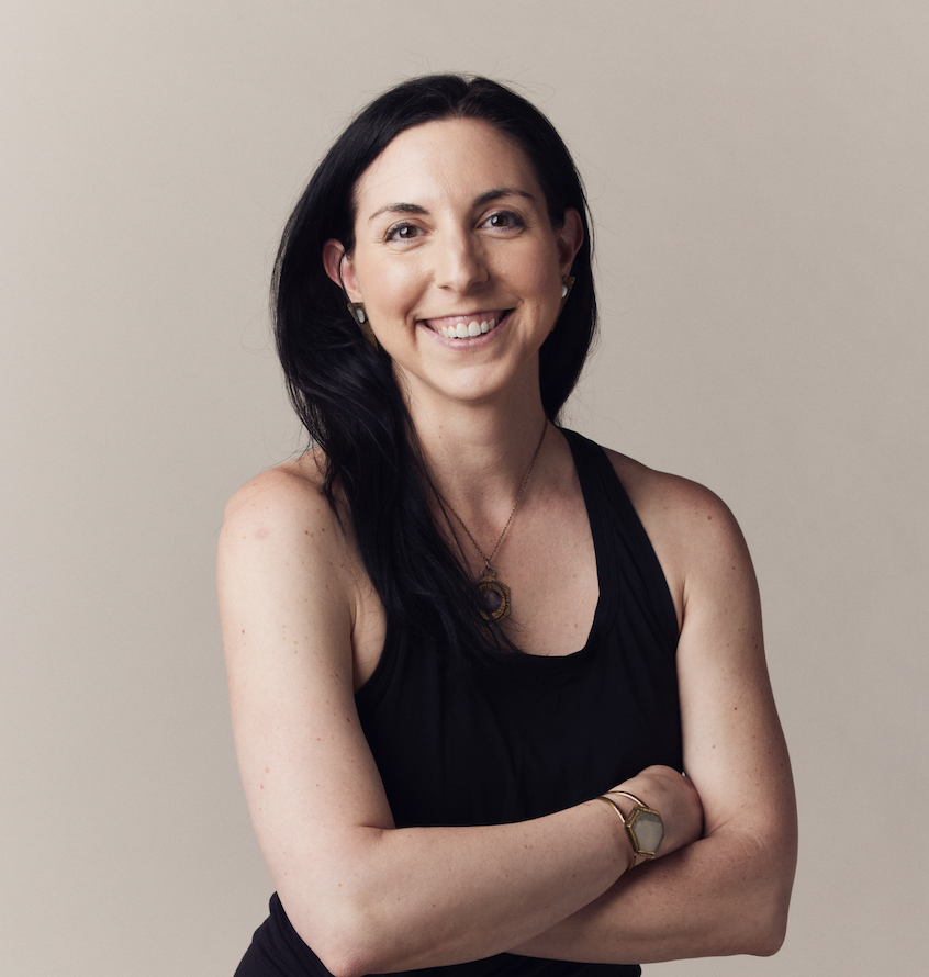 Smiling woman with long dark hair wearing a black sleeveless top, necklace, and watch against a neutral background