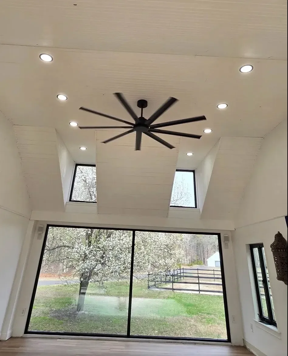 Interior shot of a room with a white ceiling featuring a black ceiling fan and recessed lighting, large sliding glass doors revealing a tree outside, and two windows on the ceiling allowing natural light in.