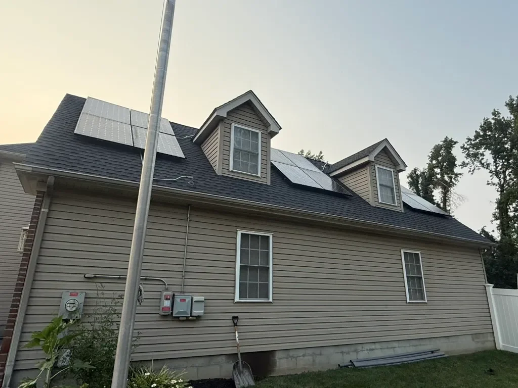 Back of a house with beige siding, three dormer windows, and solar panels on the roof, with a utility pole and some garden plants in the yard.