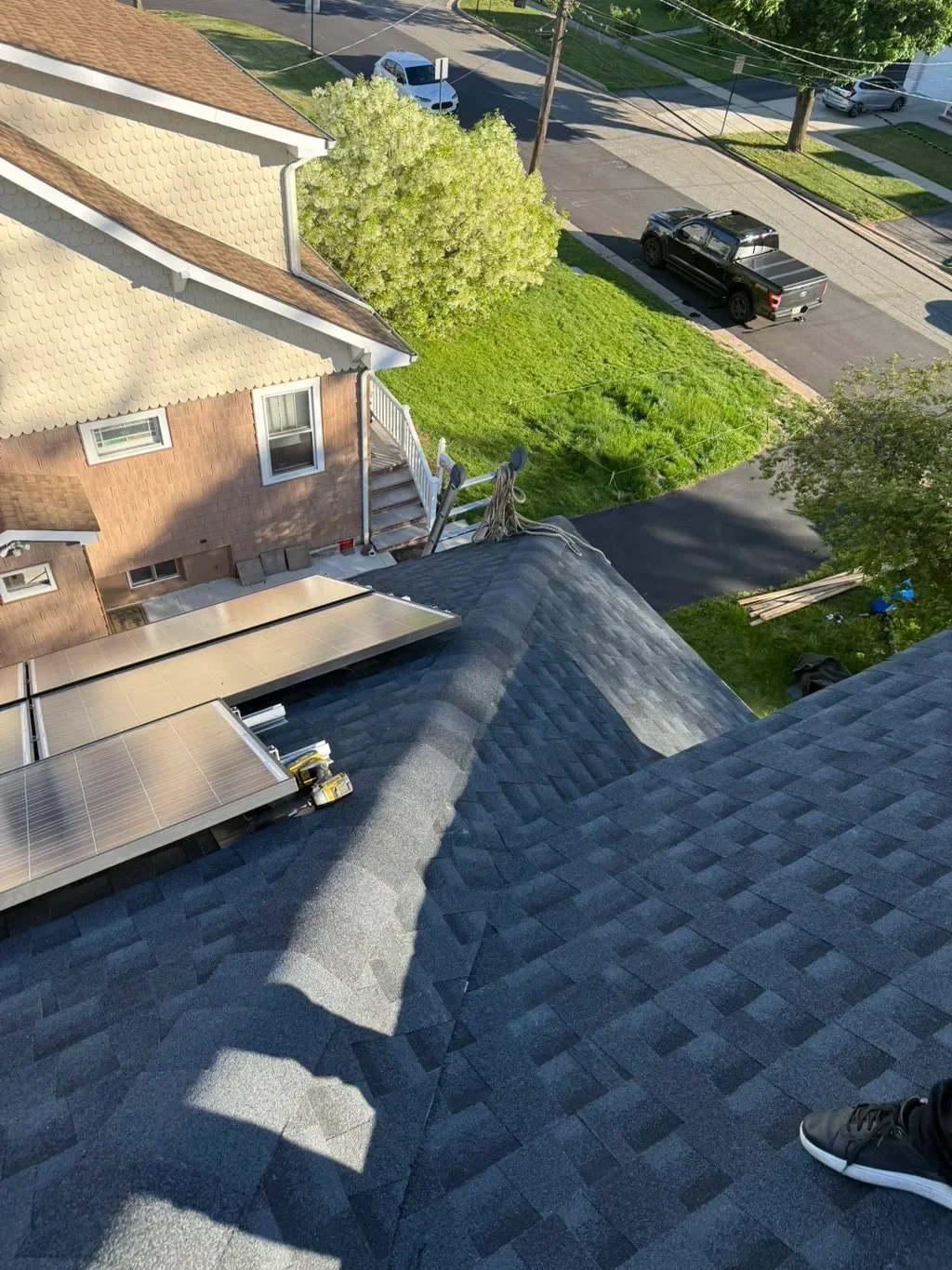 View from a rooftop showing solar panels, shingles, a sunny yard with green grass, a tree, a sidewalk, parked cars, and part of a person wearing sneakers.