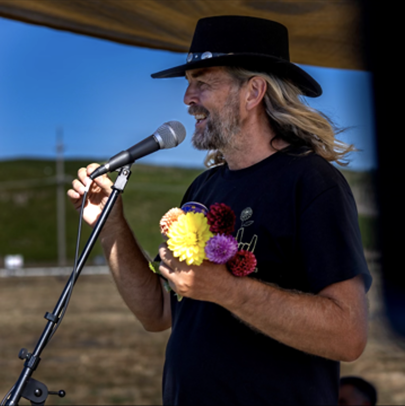 A man with long gray hair and beard, wearing a black hat and T-shirt, holding a bunch of colorful flowers, speaking into a microphone outdoors with a blue sky and green hills in the background.