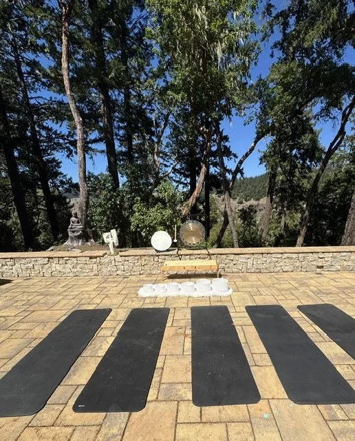 Open outdoor patio with black yoga mats laid out for a meditation session, overlooking trees and a stone wall, with meditation cushions and statues in the background.