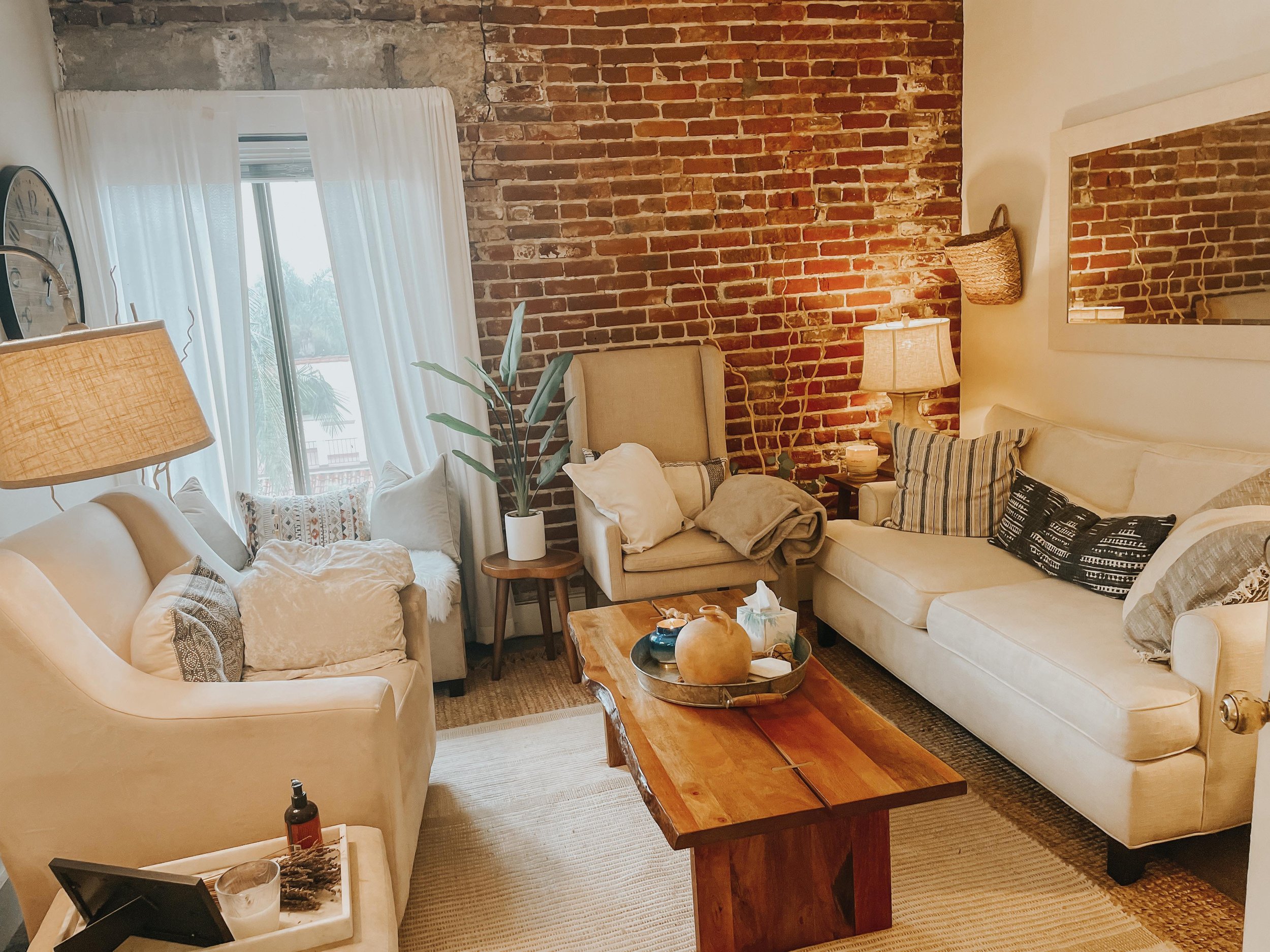 Living room with exposed brick wall, white curtains, beige sofas with patterned pillows, wooden coffee table with decorative items, a potted plant, and a mirror on the wall.