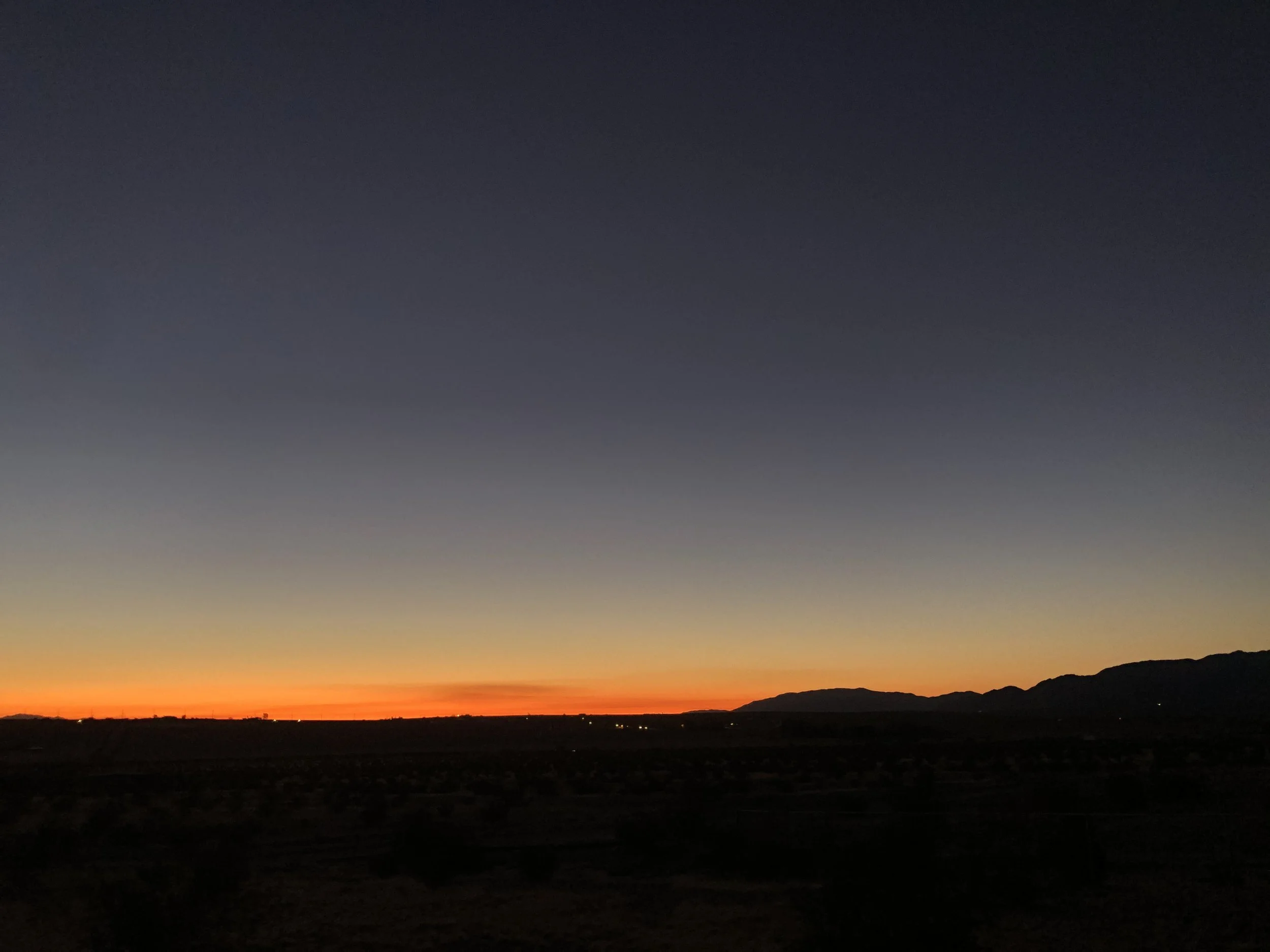 A landscape view of a sunset or sunrise with a dark silhouette of mountains in the distance, and the sky transitioning from orange near the horizon to darker blue and black higher up.