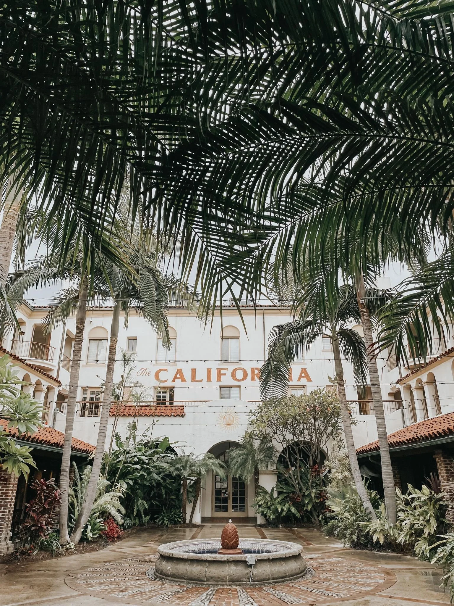 The California building with Mediterranean-style architecture, surrounded by lush tropical plants, with a fountain featuring a pineapple sculpture in the courtyard.