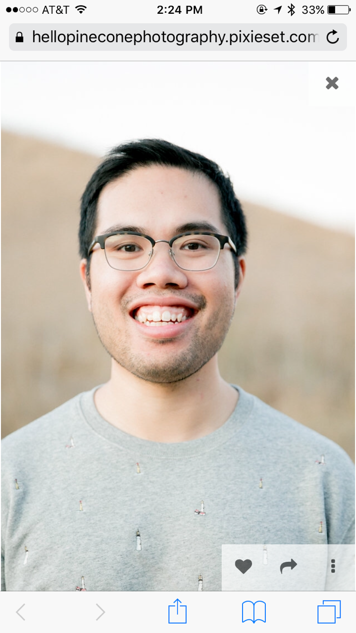 A young man with glasses smiling outdoors in front of a blurred natural background.