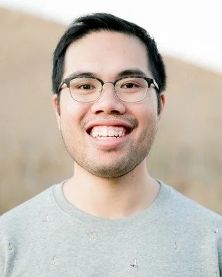 A young man with glasses smiling outdoors, with a blurred natural background.