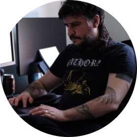 Young man with tattoos working at a computer desk in a dimly lit room.