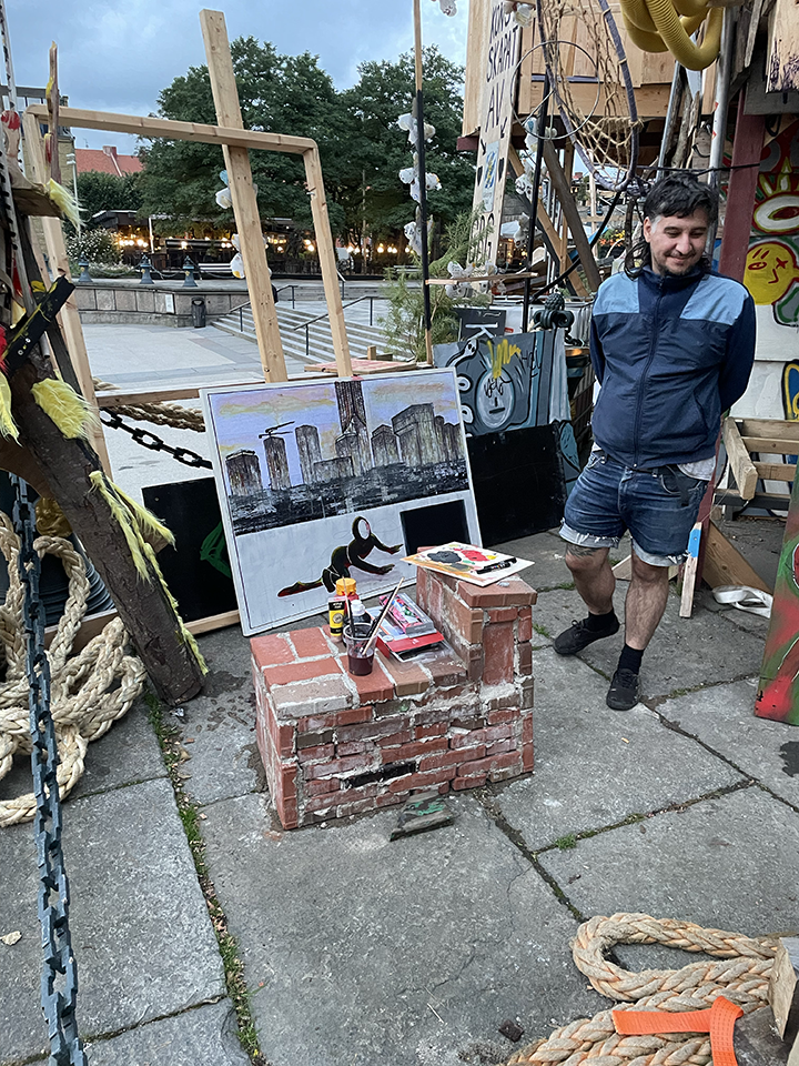 An outdoor art display showing a partially completed painting of the New York City skyline reflected in water. Art supplies are on a small brick platform in front of the painting. A man in a blue jacket and shorts stands nearby, smiling. The setting appears to be an outdoor art market or studio with other artworks and structures around.