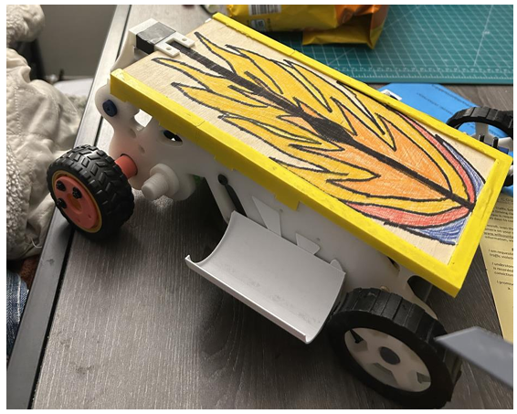 A small toy or model vehicle with black wheels, a yellow frame, and a painted flame design on the top surface, placed on a gray surface with various objects around it.