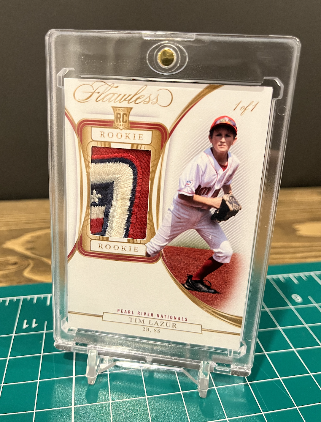 A baseball trading card featuring a young player named Tim Lazur in a batting stance, holding a baseball glove. The card includes a patch of fabric with the number 7, labeled 'Rookie', and has a picture of the player in a white uniform with a red cap. The card’s title is "Flawless" with the subtitle "Rookie" and details about the player and team, Pearl River Nationals.