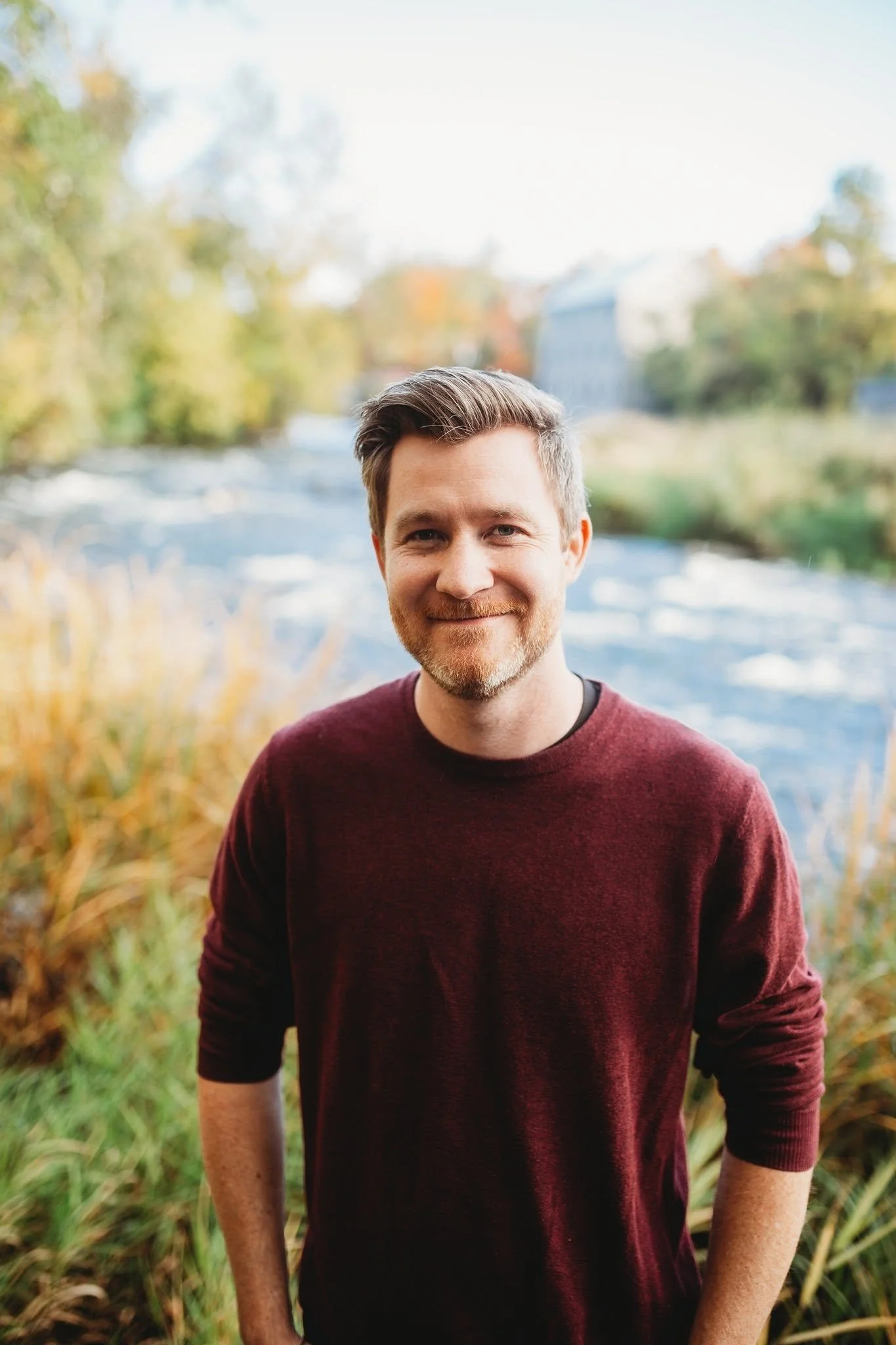 A portrait of a smiling man with brown hair and a beard, wearing a maroon long-sleeve shirt, outdoors near a river with fall foliage.