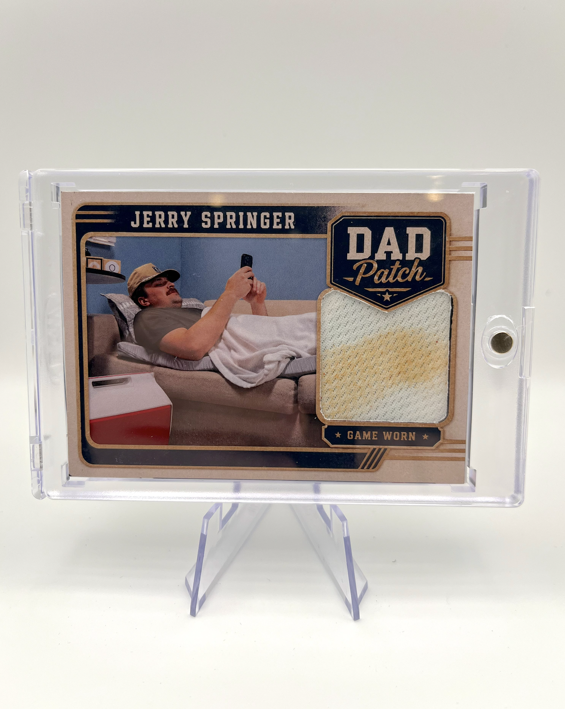 A collectible sports card of Jerry Springer with a fabric patch labeled 'Game Worn' and 'Dad Patch,' featuring an image of a man lying on a bed, using a smartphone.