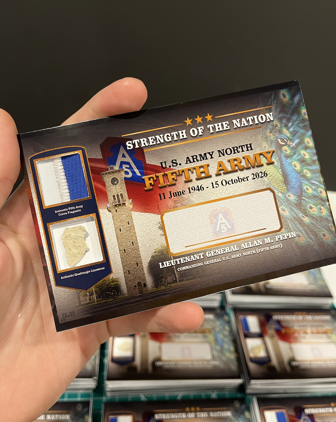 A person holding a patriotic card for the Fifth Army of the U.S. Army North, with the dates June 11, 1946, to October 15, 2026, and a peacock feather pattern in the background.