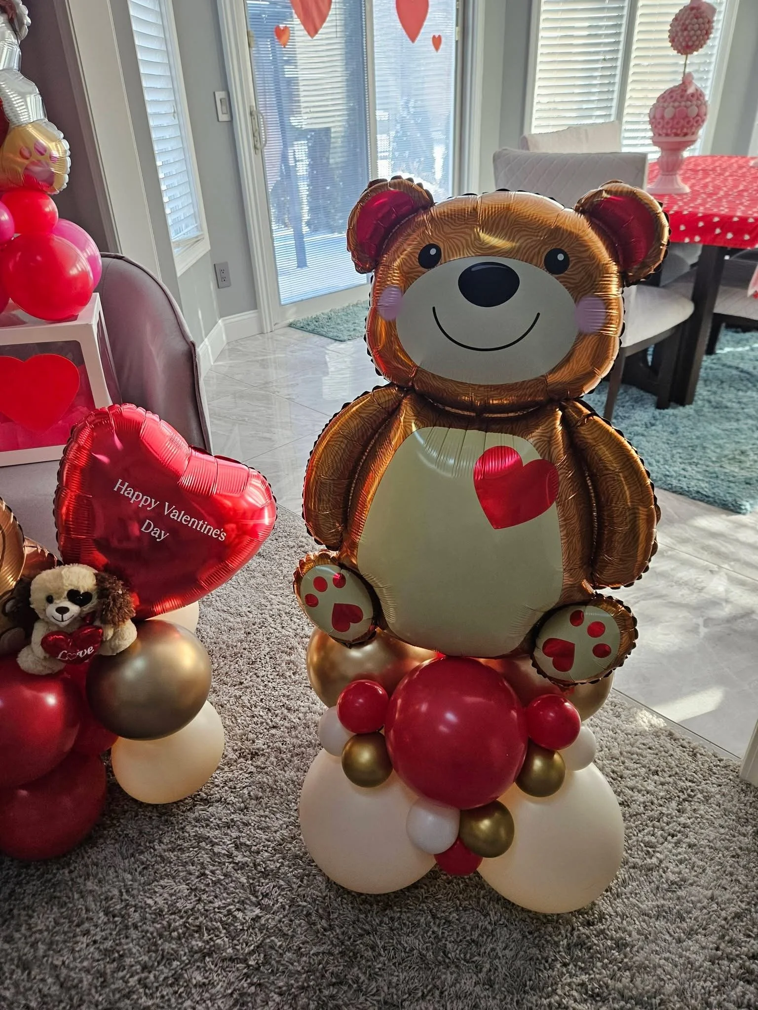 Decorative Valentine's Day balloon display featuring a large teddy bear-shaped balloon with a red heart on its chest, surrounded by pink, red, white, and gold balloons, including smaller balloons with love messages, inside a room with sliding glass doors and festive decorations.