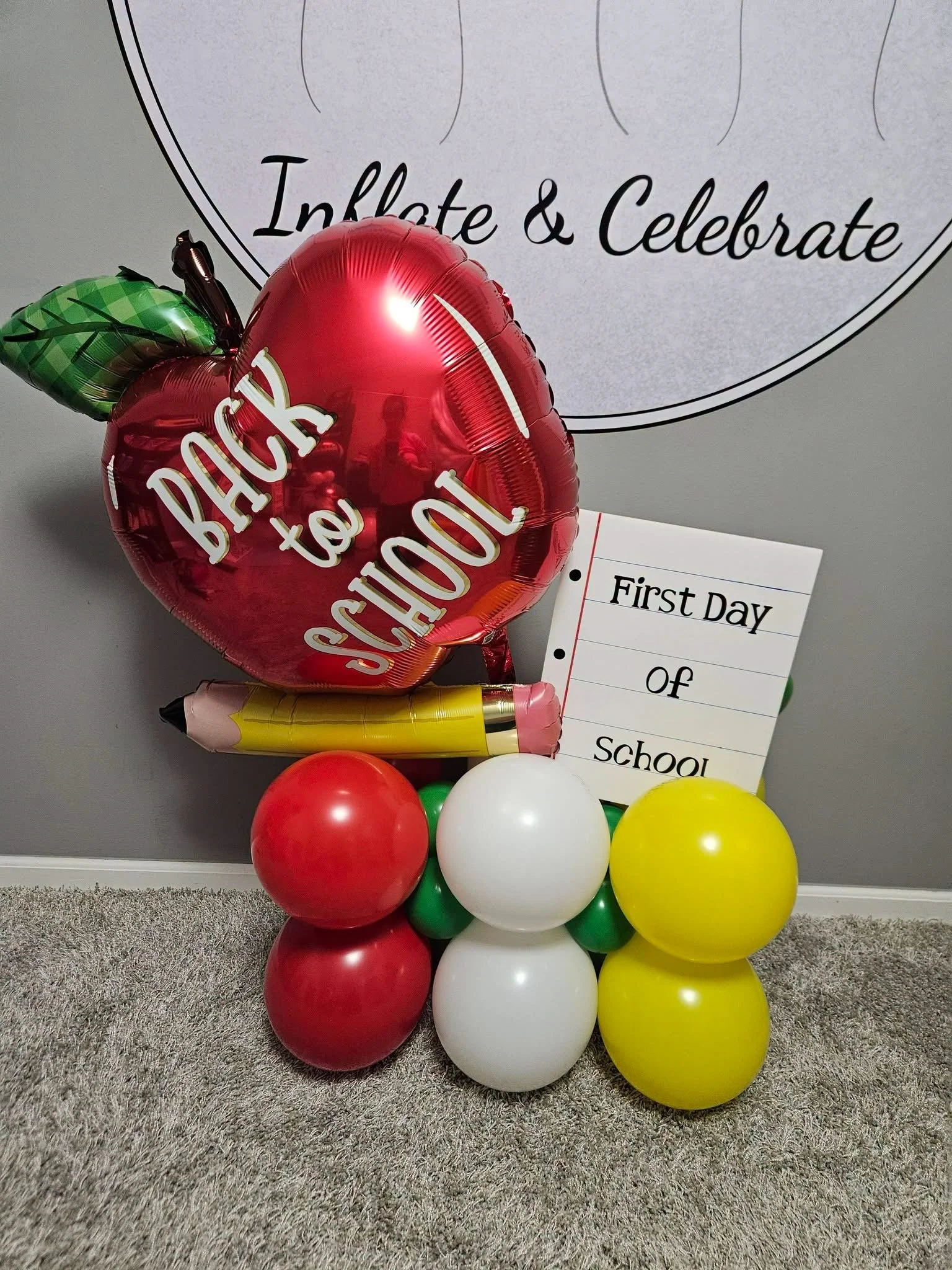 A collection of balloons, including a red apple-shaped balloon with the text 'Back to School' and a green leaf, surrounded by red, white, yellow, and green balloons, with a whiteboard that has a paper titled 'First Day of School' in the background.