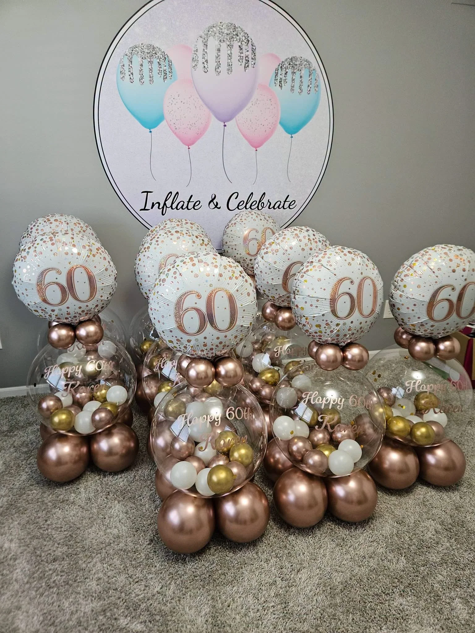 Arrangement of balloons for a 60th birthday celebration, including large rose gold and gold balloons, clear balloons with smaller balloons inside with 'Happy 60th' written on them, and a backdrop with pastel balloons and the phrase 'Inflate & Celebrate'.