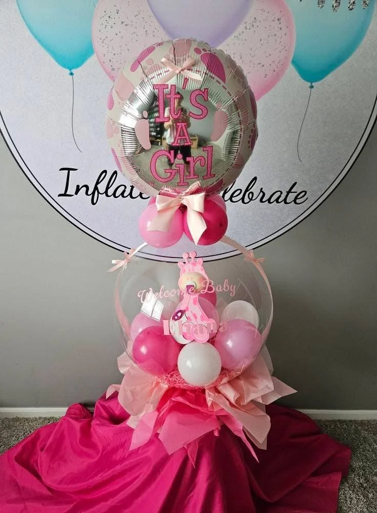 Balloon arrangement for a baby girl celebration with a large pink and silver balloon reading "It's A Girl," surrounded by pink, white, and blue balloons, and a clear balloon with a pink giraffe, baby bottle, and the words "Welcome Baby" inside, all on pink and purple fabric.