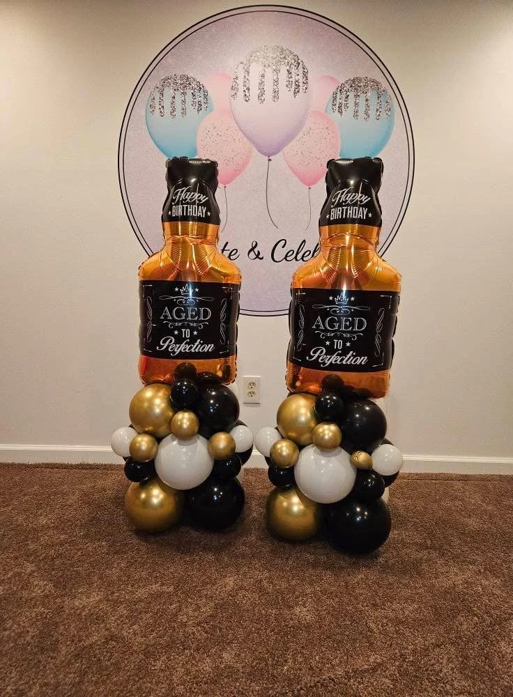 Balloon photo display with large champagne bottle-shaped balloons that say "Happy Birthday" and "Aged to Perfection," surrounded by clusters of black, white, and gold balloons, set against a wall with a birthday poster featuring pink and blue balloons.