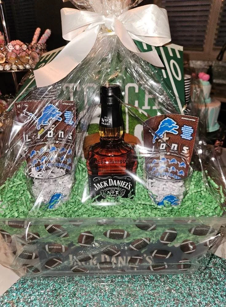 Gift basket with Jack Daniel's whiskey, branded shot glasses, and Detroit Lions memorabilia, wrapped in clear cellophane with a white ribbon on top.