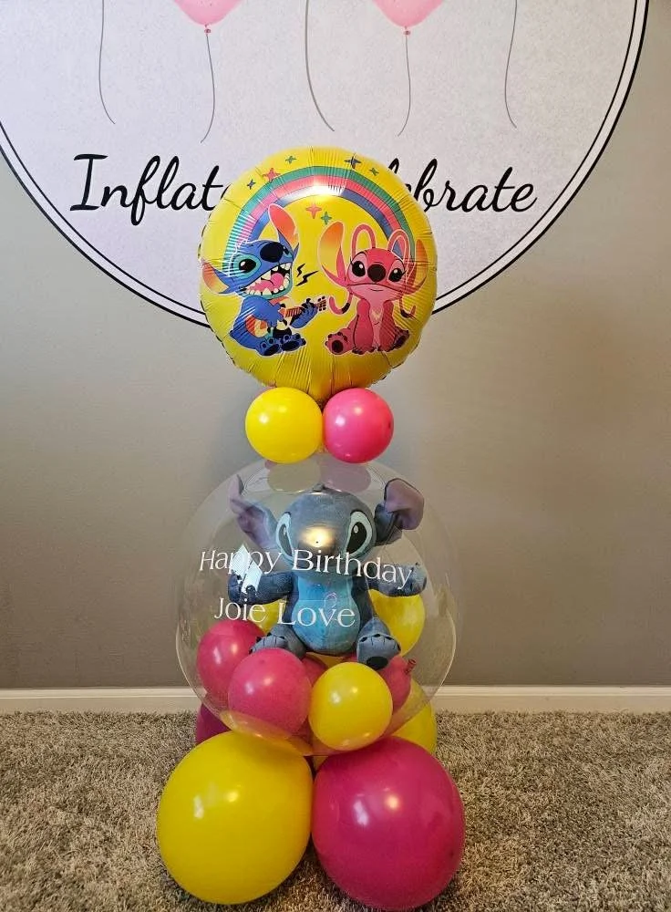 Colorful birthday balloon arrangement with a large character balloon at the top, featuring two animated animal characters, a pink and a blue one. Below is a clear balloon with a stuffed elephant inside, with white text saying 'Happy Birthday Joie Love'. The base consists of yellow and pink balloons on a carpeted floor.