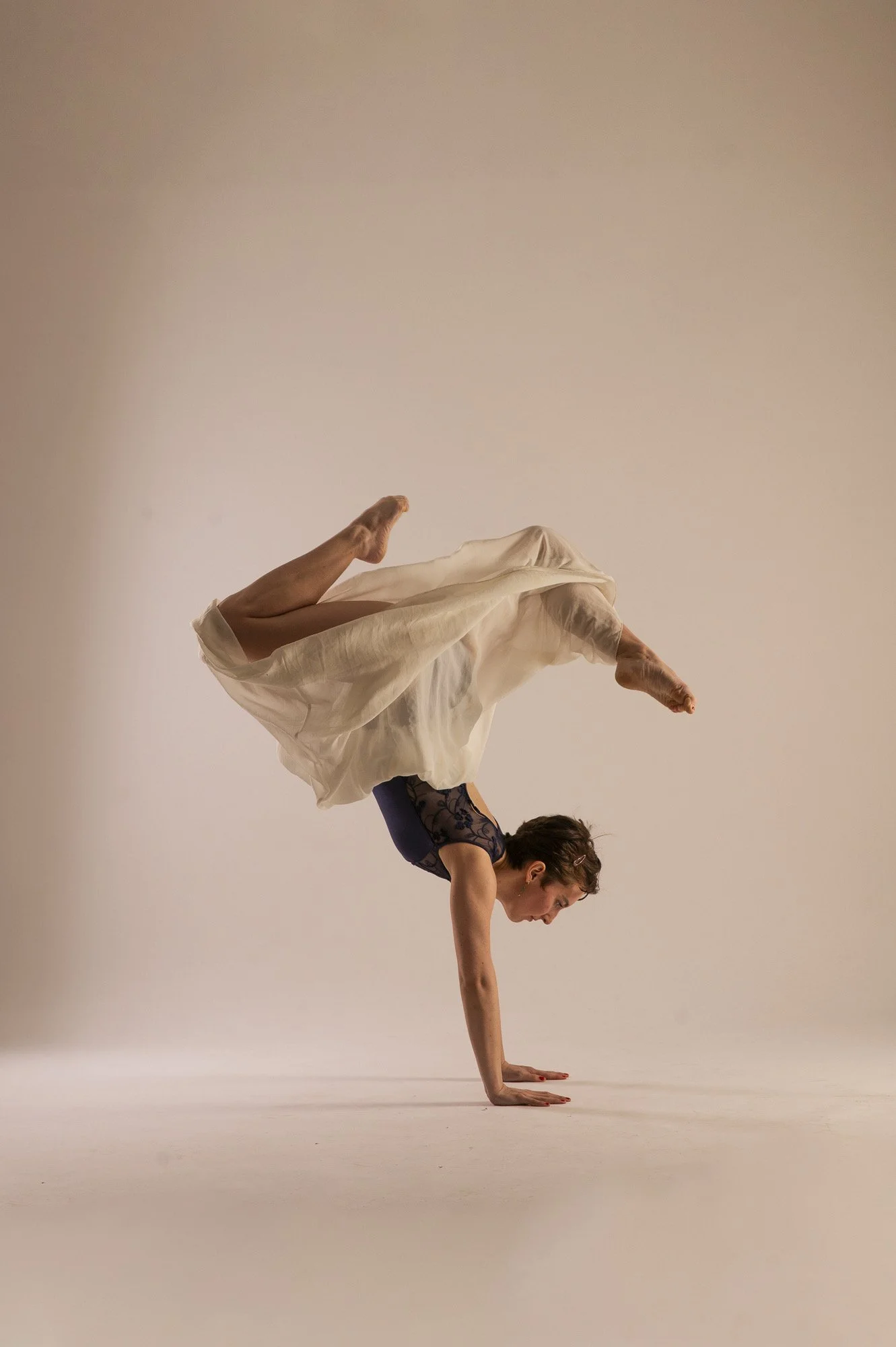 A woman performing a yoga pose, balancing on her hands with her legs raised above her head, wearing a black crop top and flowing white skirt.