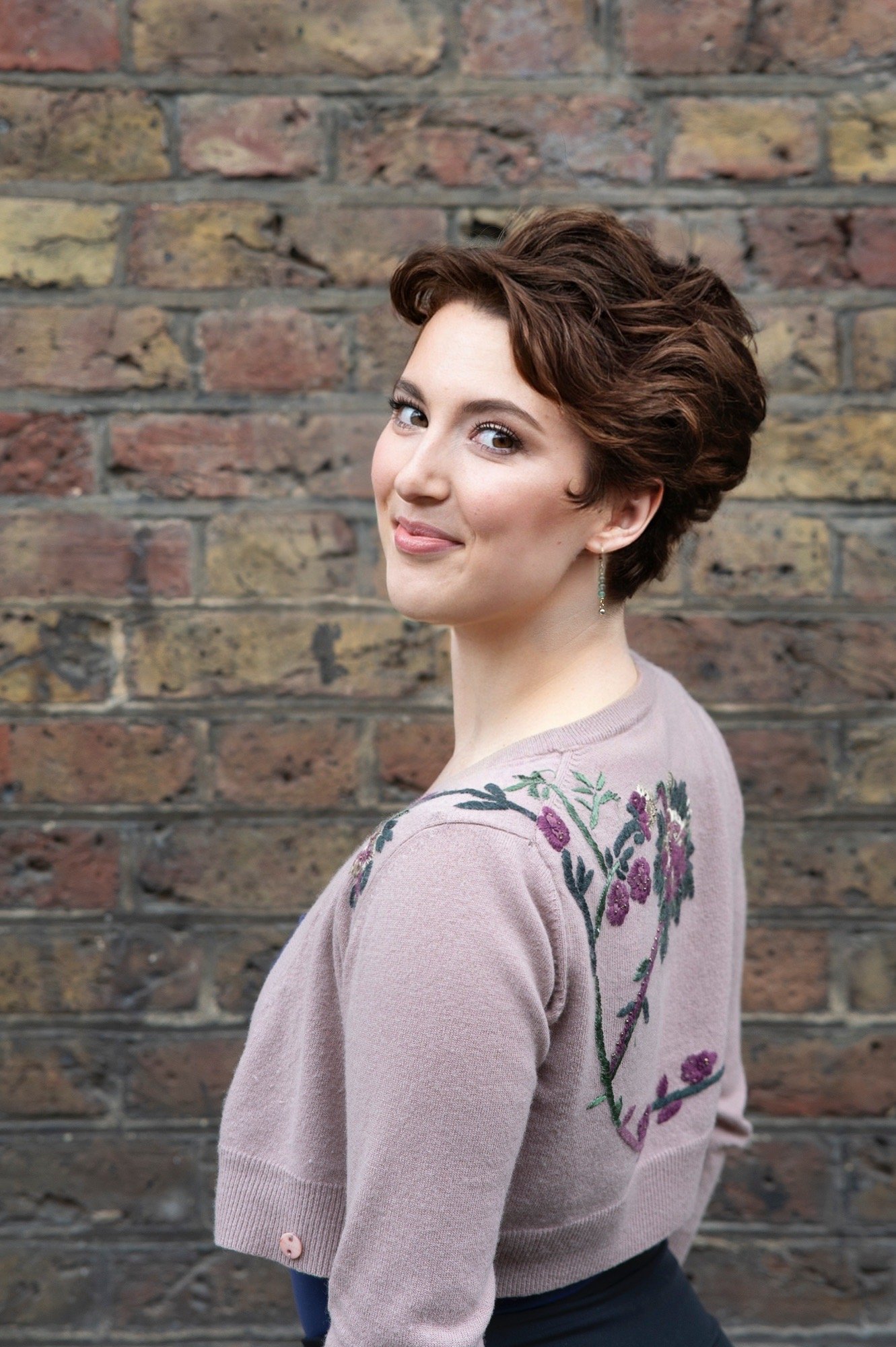 Portrait of a woman with short curly brown hair, wearing a light pink embroidered sweater, standing against a brick wall, smiling slightly and looking at the camera.