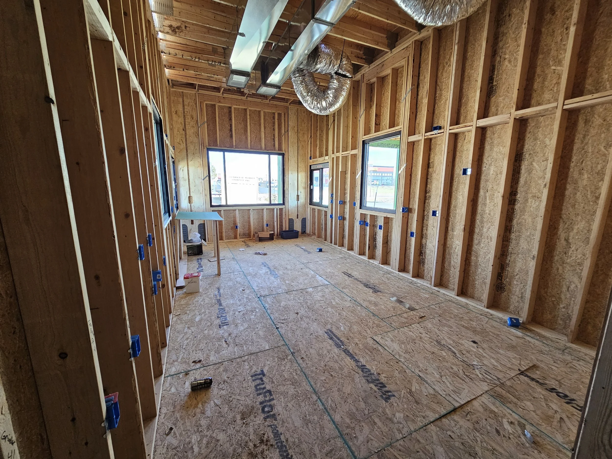 Interior of a house under construction with exposed wooden framing, ceiling ductwork, and open windows, showing a view outside.