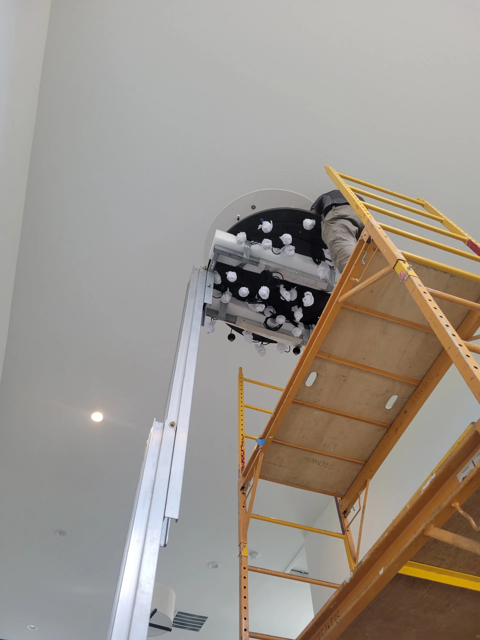 Worker on orange scaffolding installing or repairing ceiling lights in a white room.