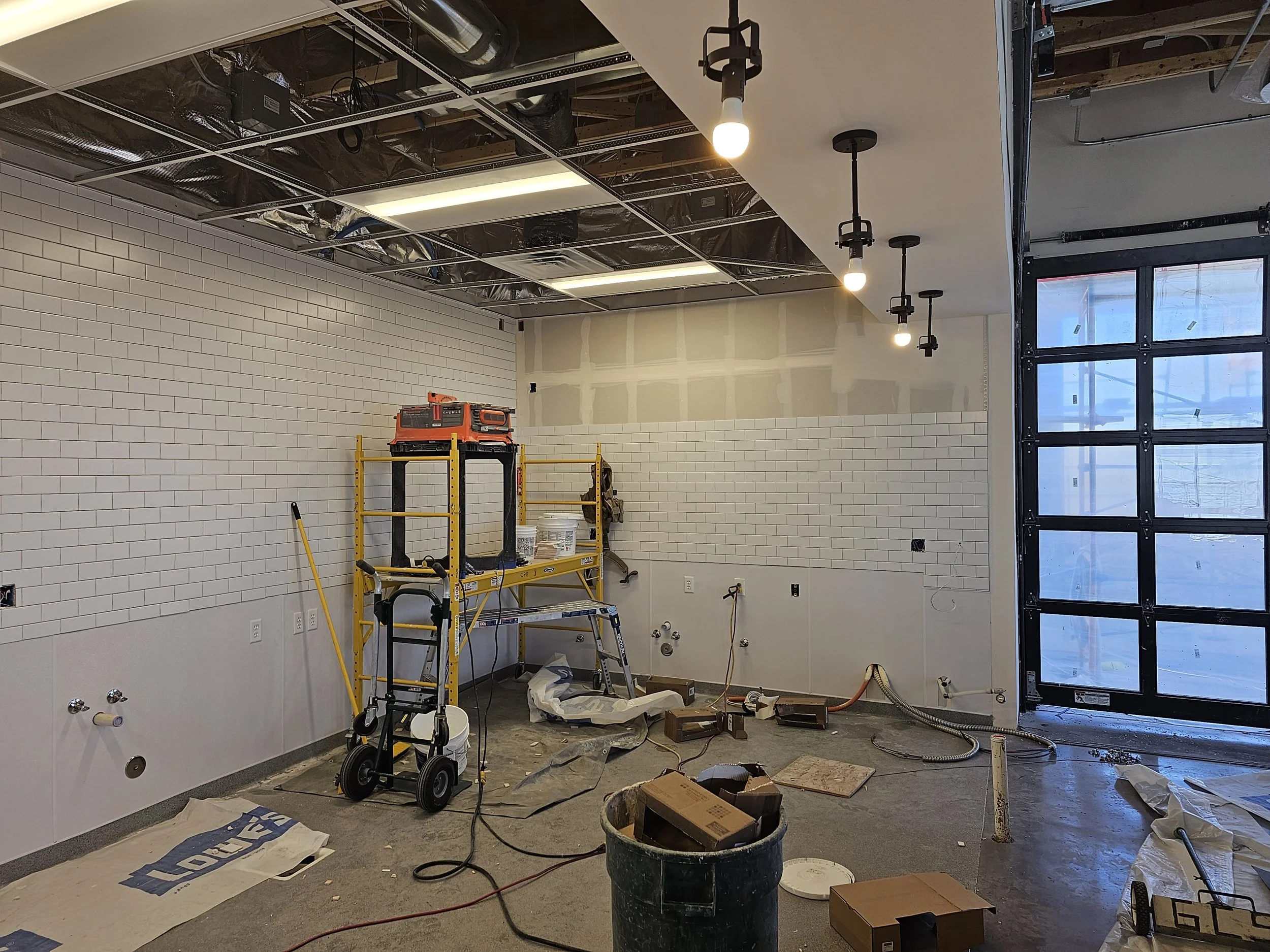 Construction site inside a building with unfinished white tiled walls, tools, and equipment scattered around. The ceiling is partly open with visible ductwork and lighting fixtures. There are large windows on one side.