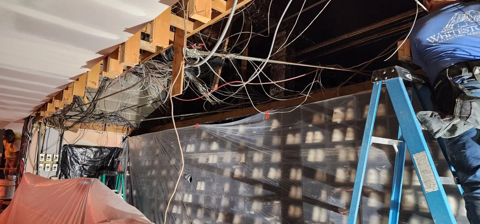 Interior of a building under renovation with exposed electrical wiring, plastic sheeting, and construction workers.