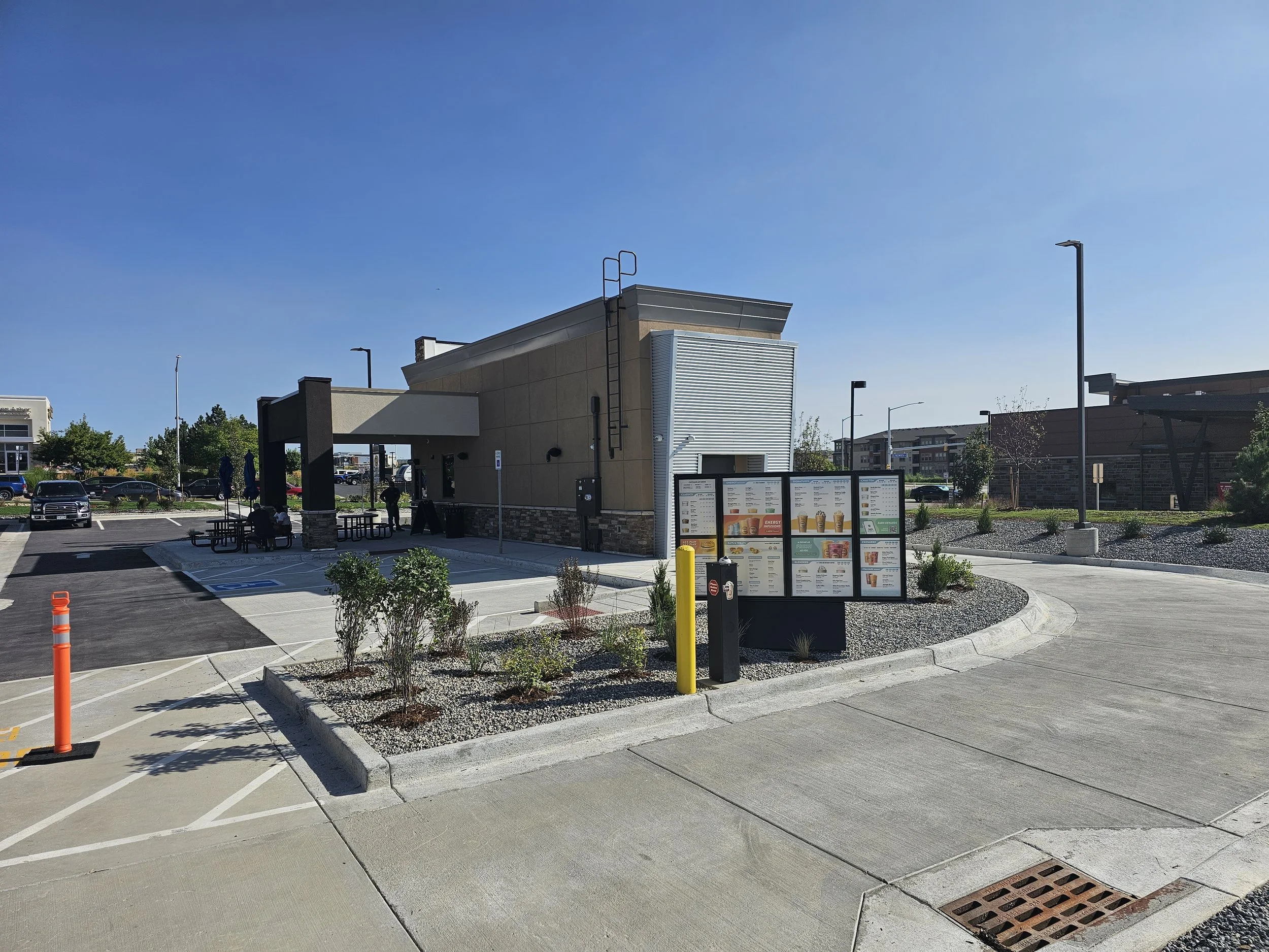 A small strip mall or fast food restaurant with outdoor seating and a parking lot in front, under a clear blue sky.