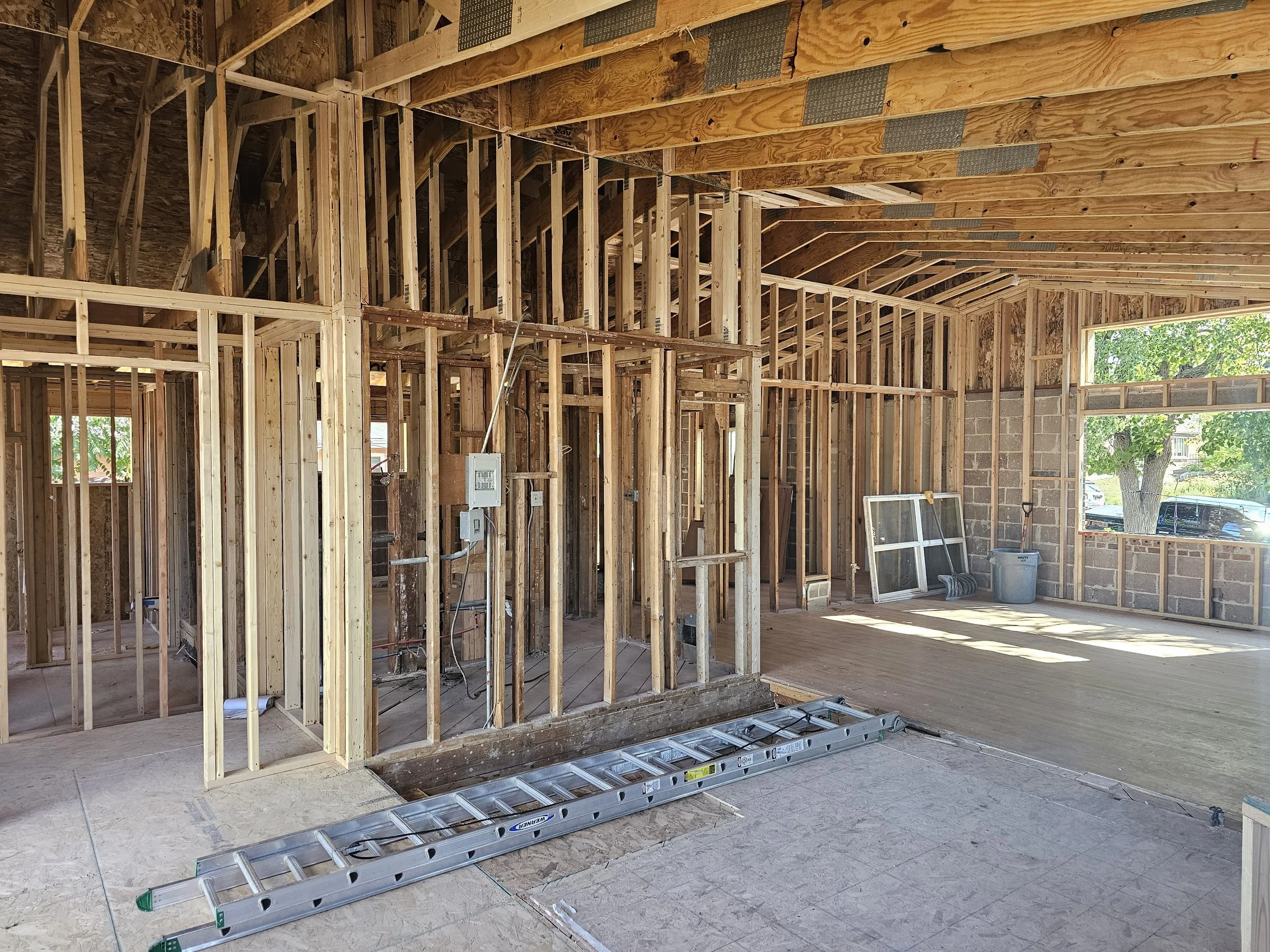 Interior view of a house under construction showing exposed wooden framing, electrical wiring, and a large window with a view of trees outside.